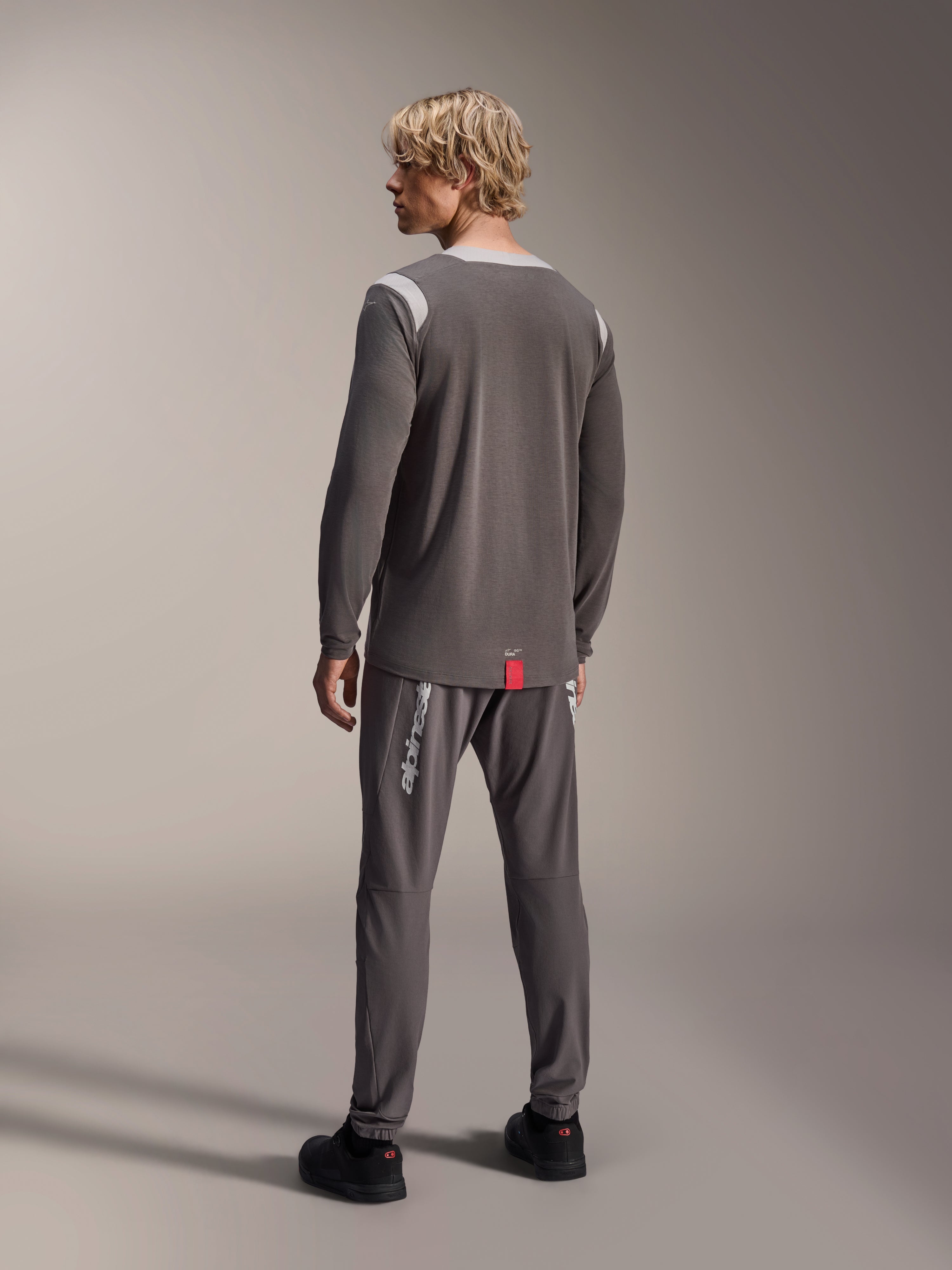 Mandlig model iført Alpinestars A-Dura Dri Wool langærmet trøje, mørkegrå med lysegrå mesh-skulderpaneler, kombineret med matchende mørkegrå cykelbukser, set bagfra med fokus på den forlængede ryg, mountainbike-beklædning
