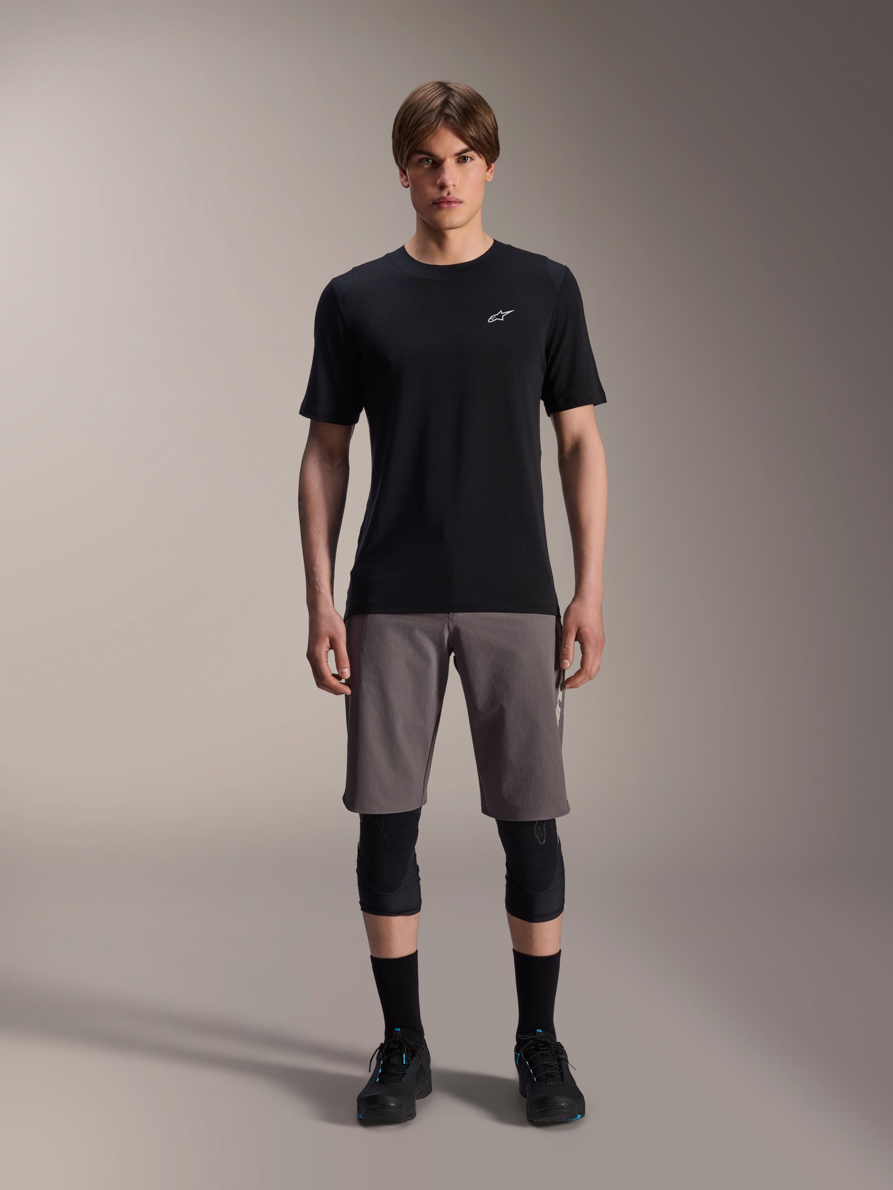 Alpinestars A-Dura Dri Wool Short Sleeve Jersey, Black, worn by a model with grey mountain bike shorts, black knee guards, and black cycling shoes, featuring a minimalist design with a small white Astars logo on the chest, functional performance MTB apparel for trail riding.