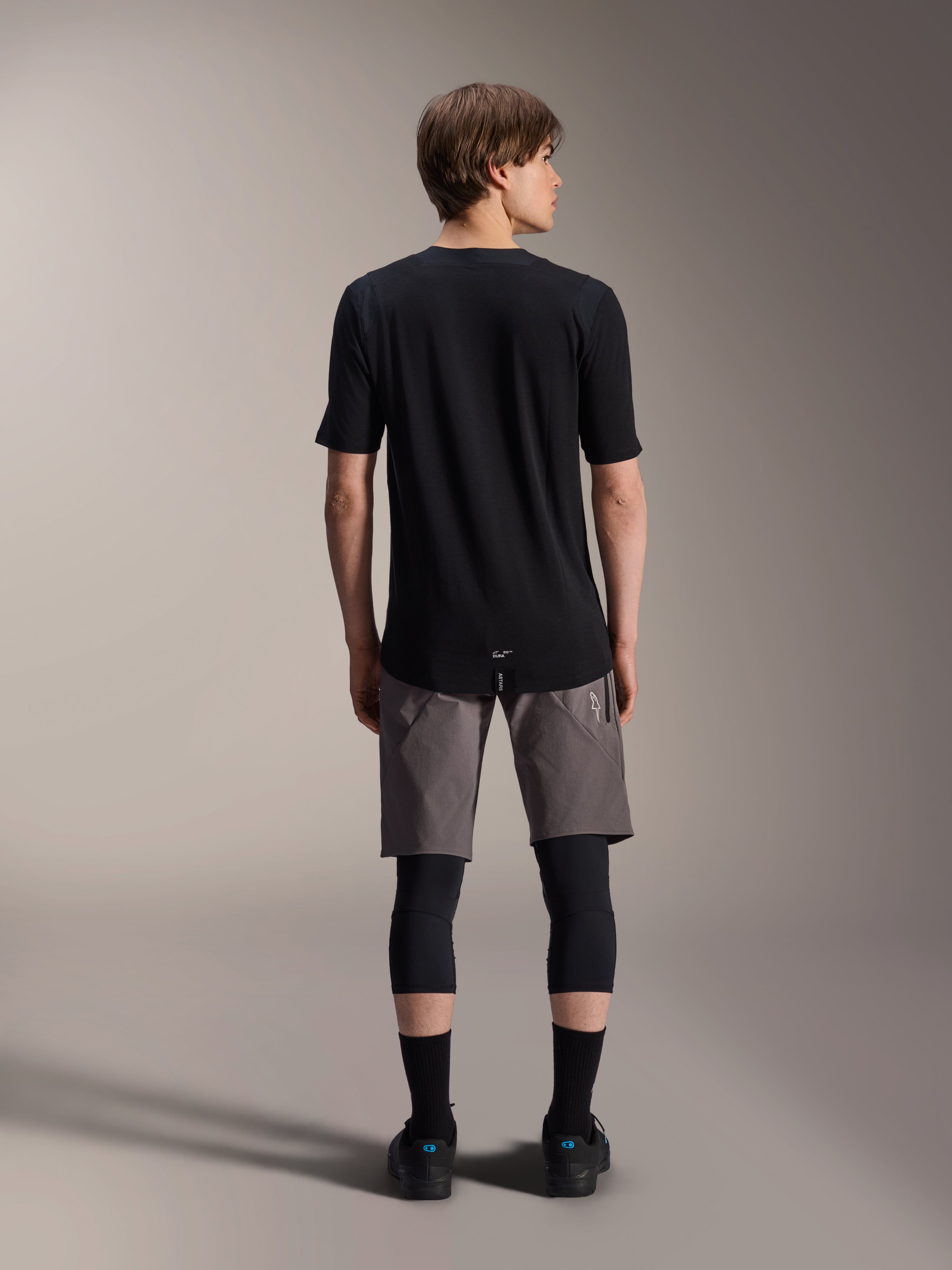 Alpinestars A-Dura Elite Shorts, Dark Gray mountain bike shorts, back view, worn with a black short-sleeve jersey, knee pads, and black socks, technical cycling gear for off-road riding