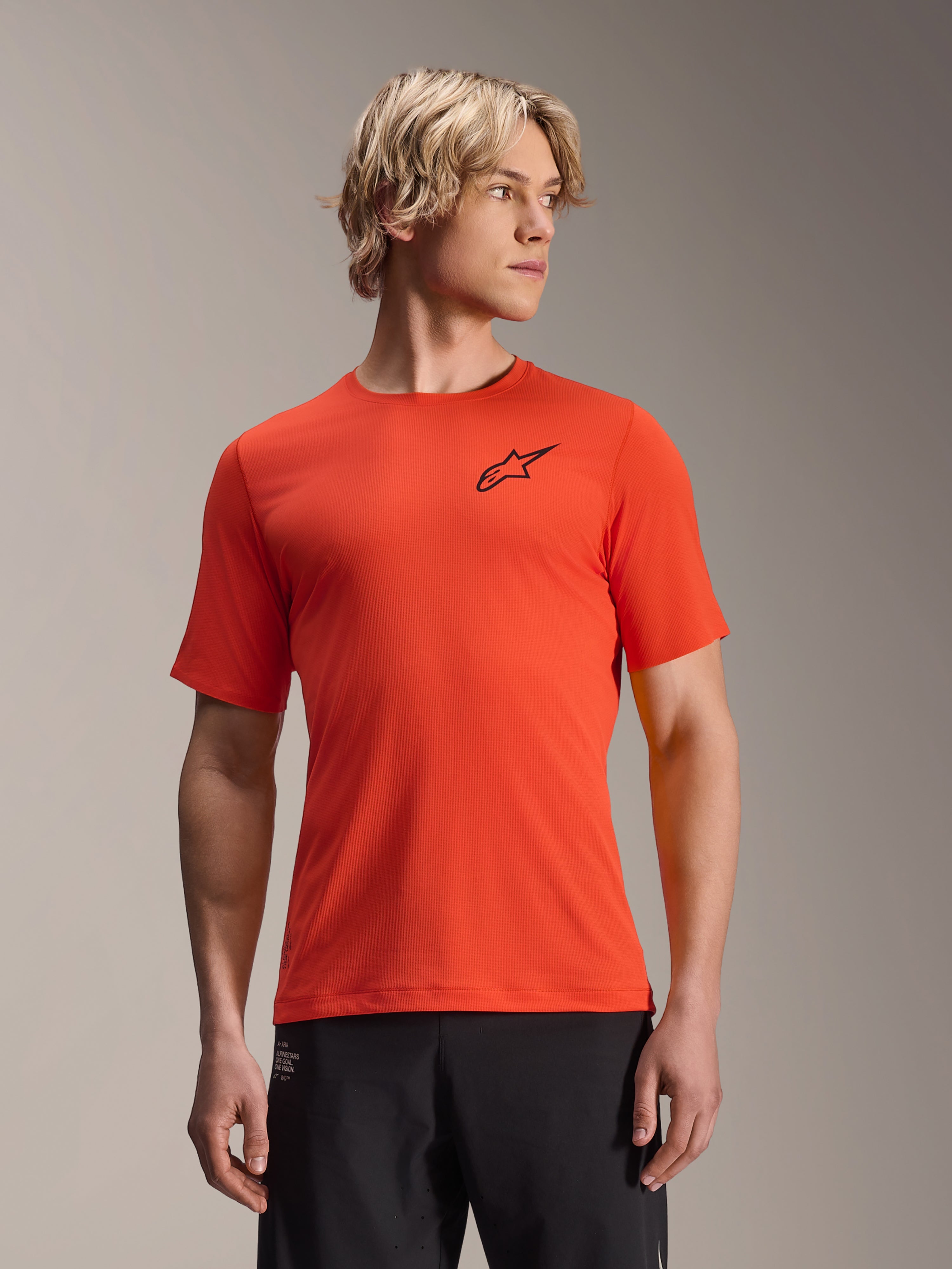 Alpinestars A-Aria Astar kortærmet trøje, Spicy Orange med sort stjerne-logo, MTB mountainbike-trøje til mænd, performance fit, båret af mandlig model med lyst hår og sorte bukser