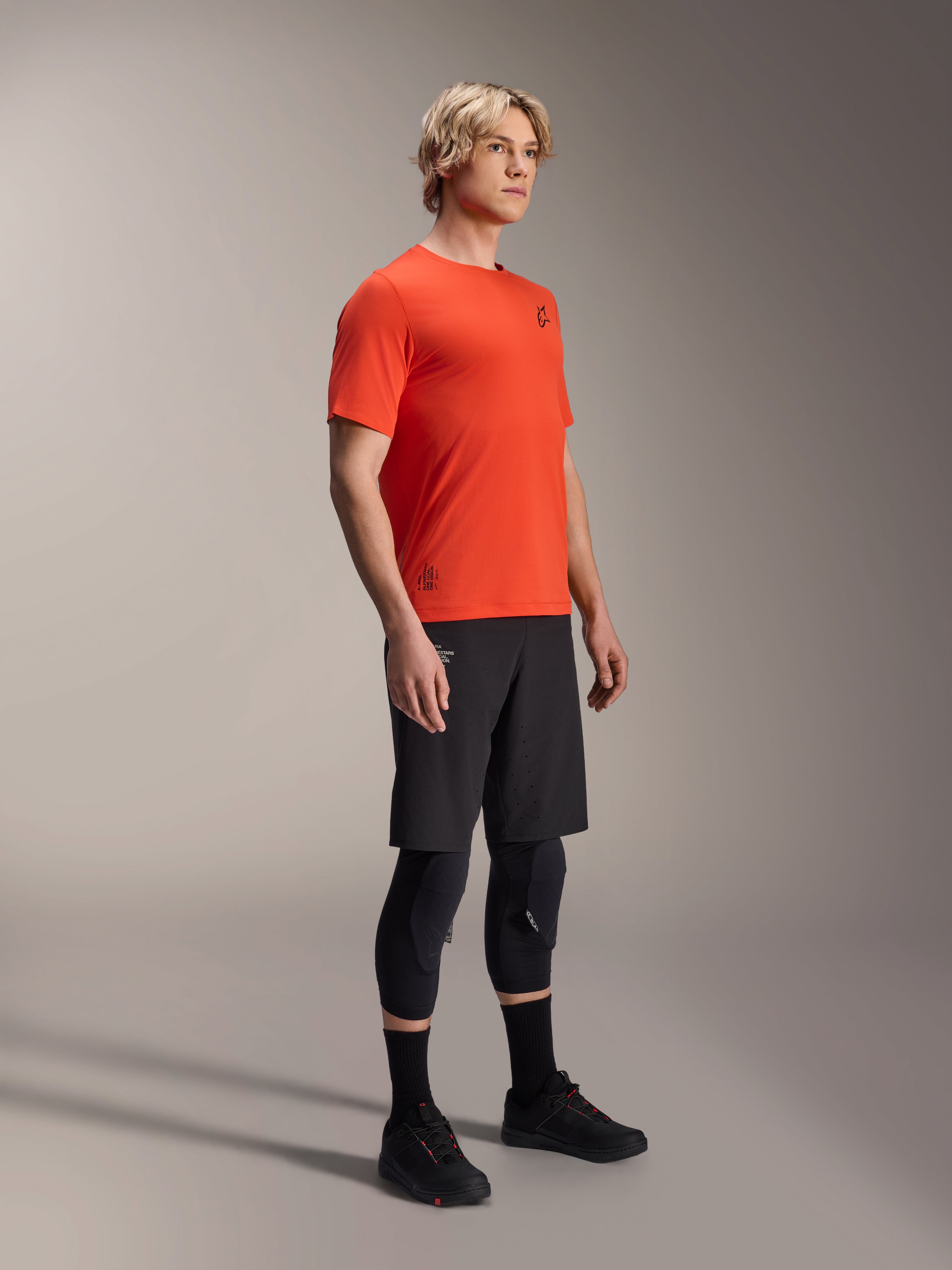 Alpinestars A-Aria Astar kortærmet trøje i Spicy Orange, båret af en model med sorte MTB-shorts og knæbeskyttelse, letvægts åndbart mountainbike-tøj, levende orange med minimal sort branding