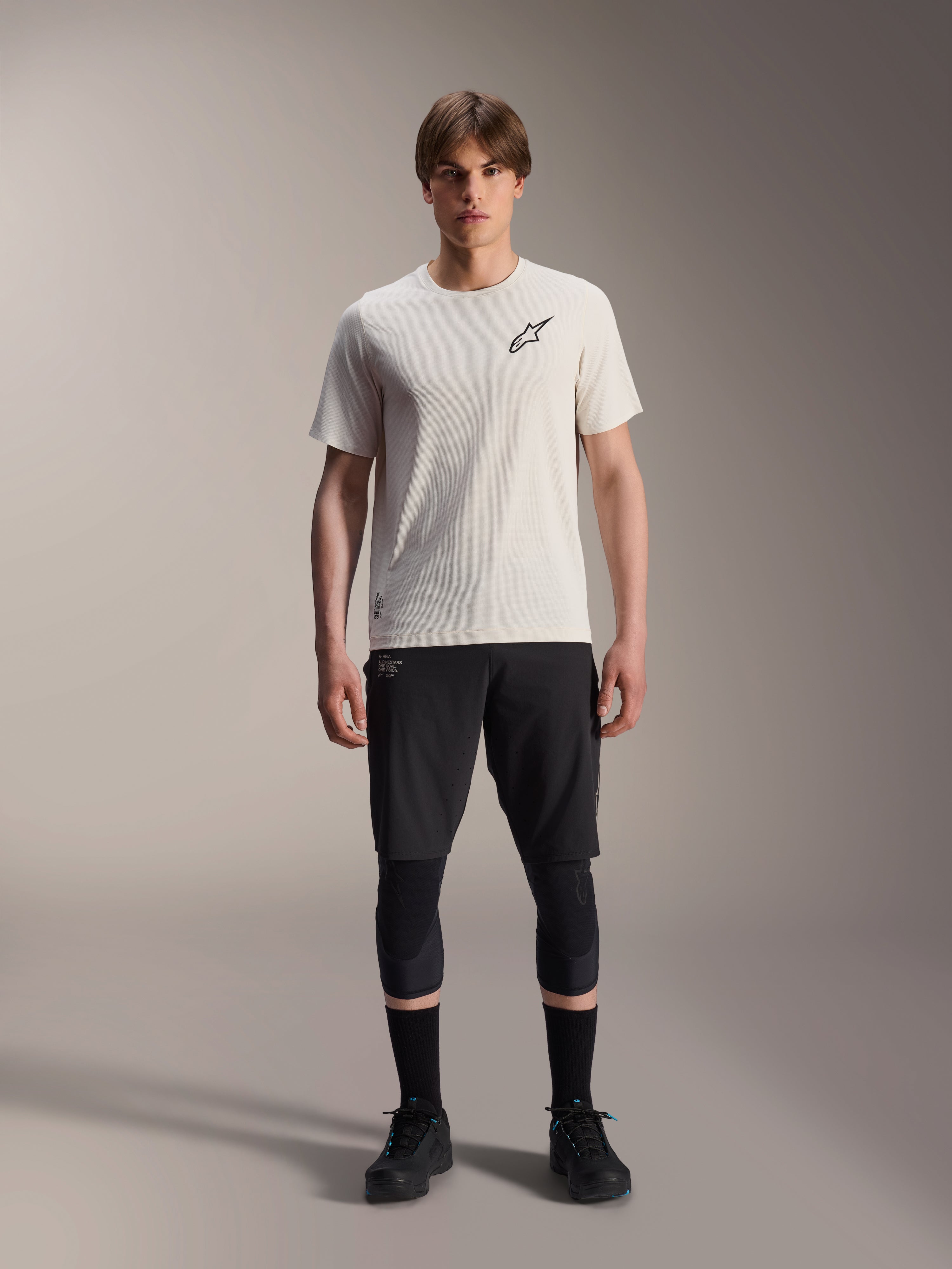 Mandlig model iført Alpinestars A-Aria Astar kortærmet trøje i Ecru, kombineret med sorte MTB-shorts, knæbeskyttere og sorte cykelsko, med et minimalistisk sort stjernelogo på brystet, designet til off-road terrænkørsel