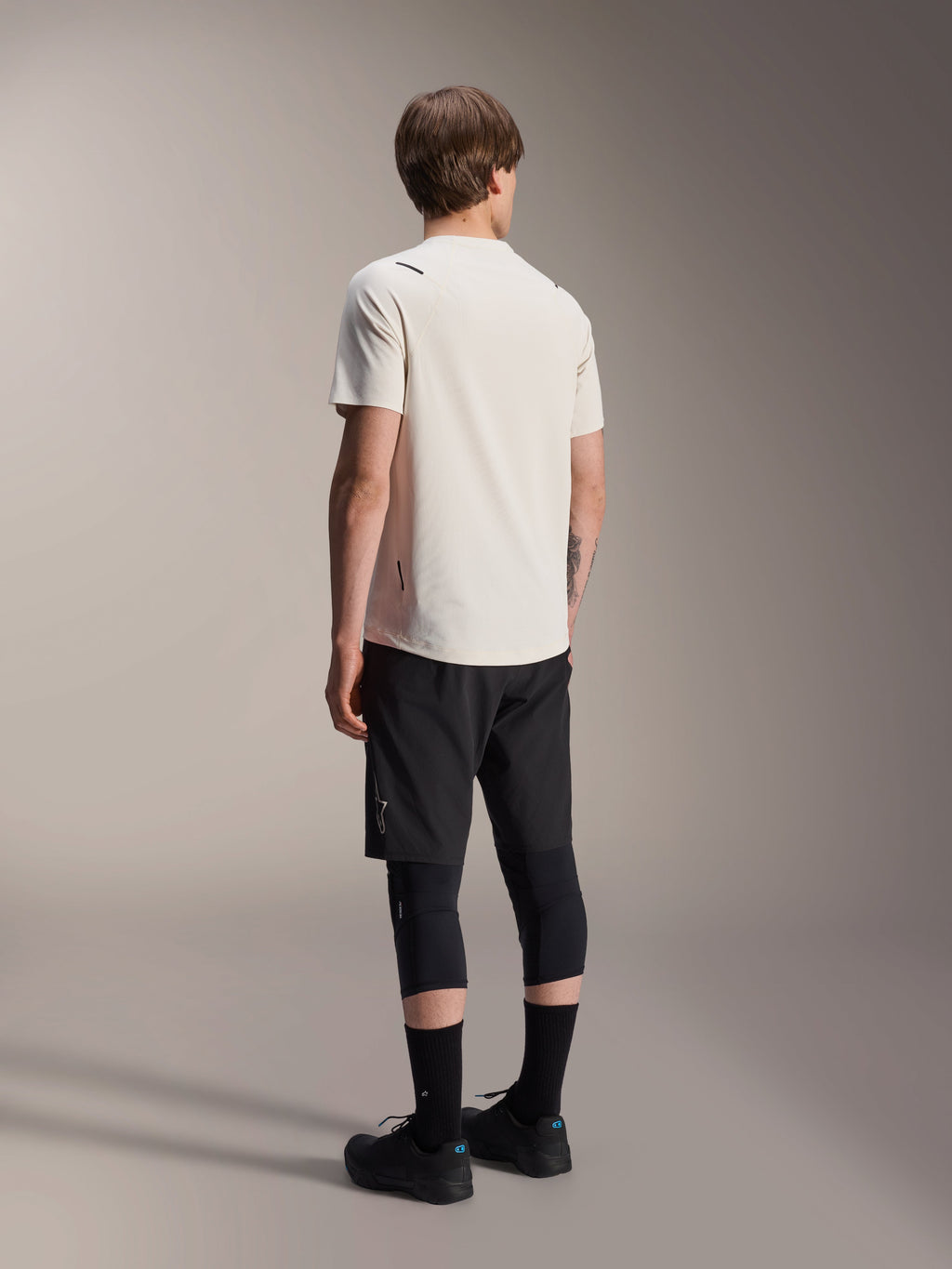 Alpinestars A-Aria Astar Jersey, kortærmet MTB-trøje, Ecru, set bagfra, viser performance-pasformen og det længere bagfald, sammensat med sorte mountainbike-shorts, knæbeskyttere og sorte cykelsko til off-road trail-sessioner