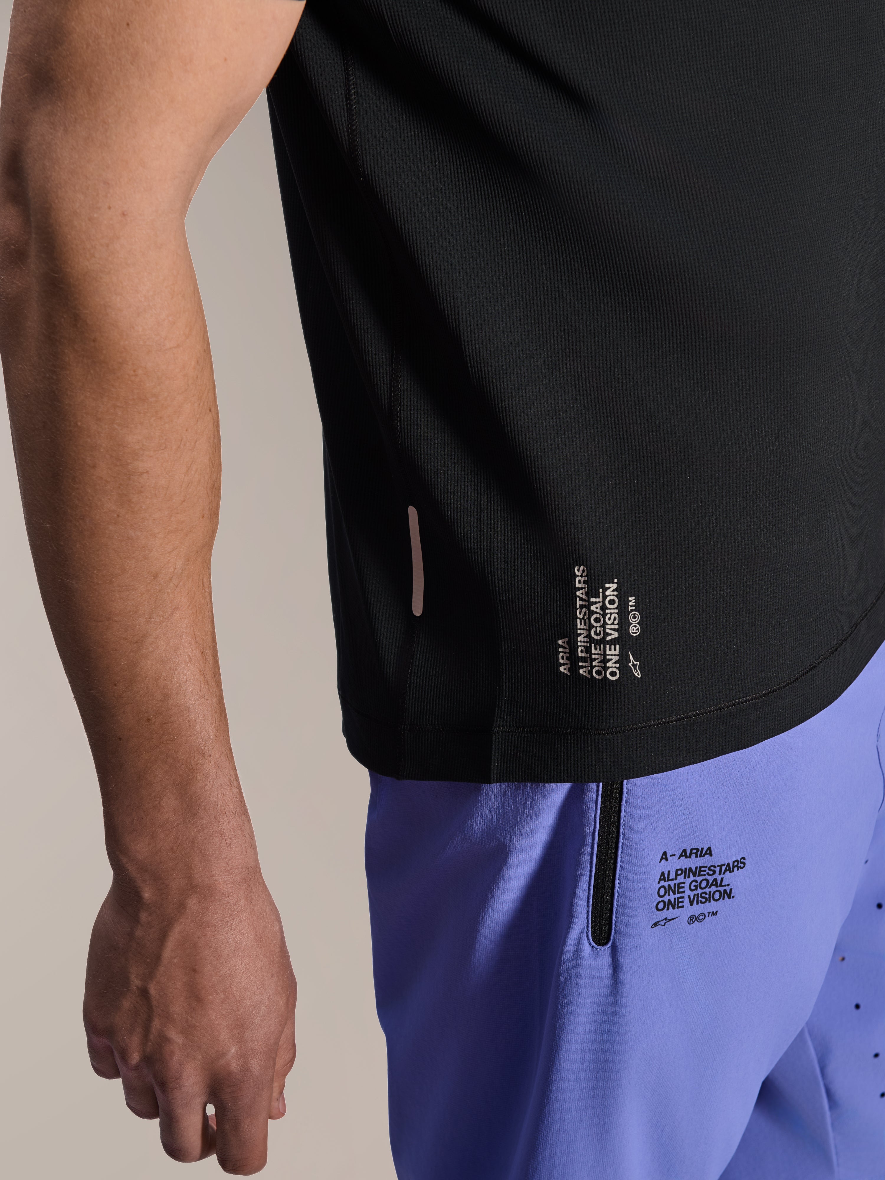 Alpinestars A-Aria Switch Kortærmet Jersey, sort MTB-trøje, nærbillede af sidekant med hvid 'ONE GOAL. ONE VISION.'-branding, båret med lyslilla cykelshorts med matchende logoer