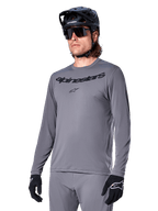 Alpinestars A-Dura Rocker Long Sleeve Jersey, Gray MTB Mountain Biking Jersey, worn with black helmet and gloves, featuring black wordmark on the chest, sporty cycling apparel for off-road riding, plain background