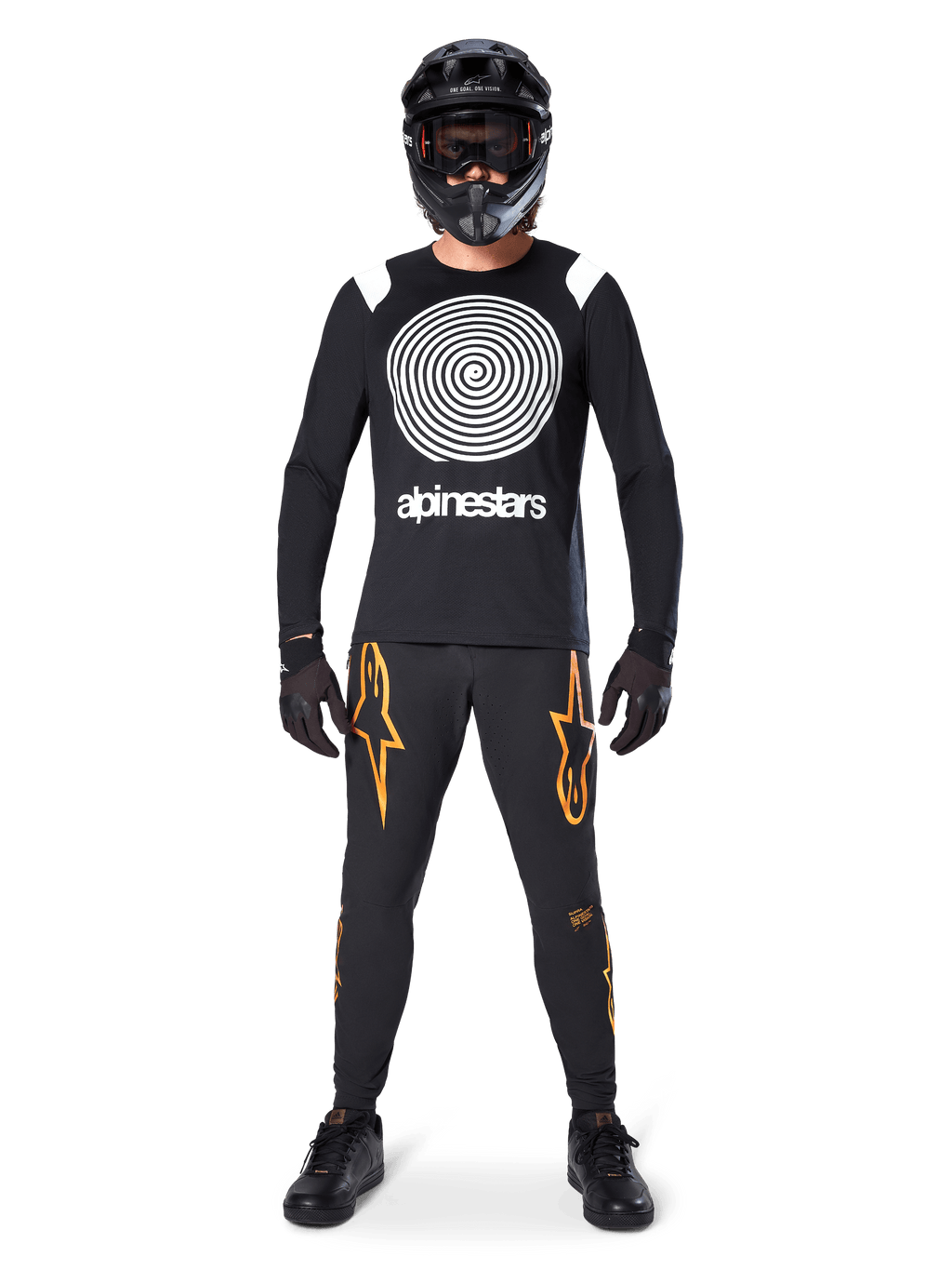 Mandlig model iført komplet mountainbike-udstyr, Alpinestars A-Supra Oscar langærmet trøje i sort med hvid spiralgrafik på brystet, sorte bukser med orange stjernelogoer, sort full-face hjelm og goggles, sorte handsker og sko, koordineret outfit til MTB gravity-kørsel