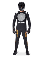 Mandlig model iført komplet mountainbike-udstyr, Alpinestars A-Supra Oscar langærmet trøje i sort med hvid spiralgrafik på brystet, sorte bukser med orange stjernelogoer, sort full-face hjelm og goggles, sorte handsker og sko, koordineret outfit til MTB gravity-kørsel