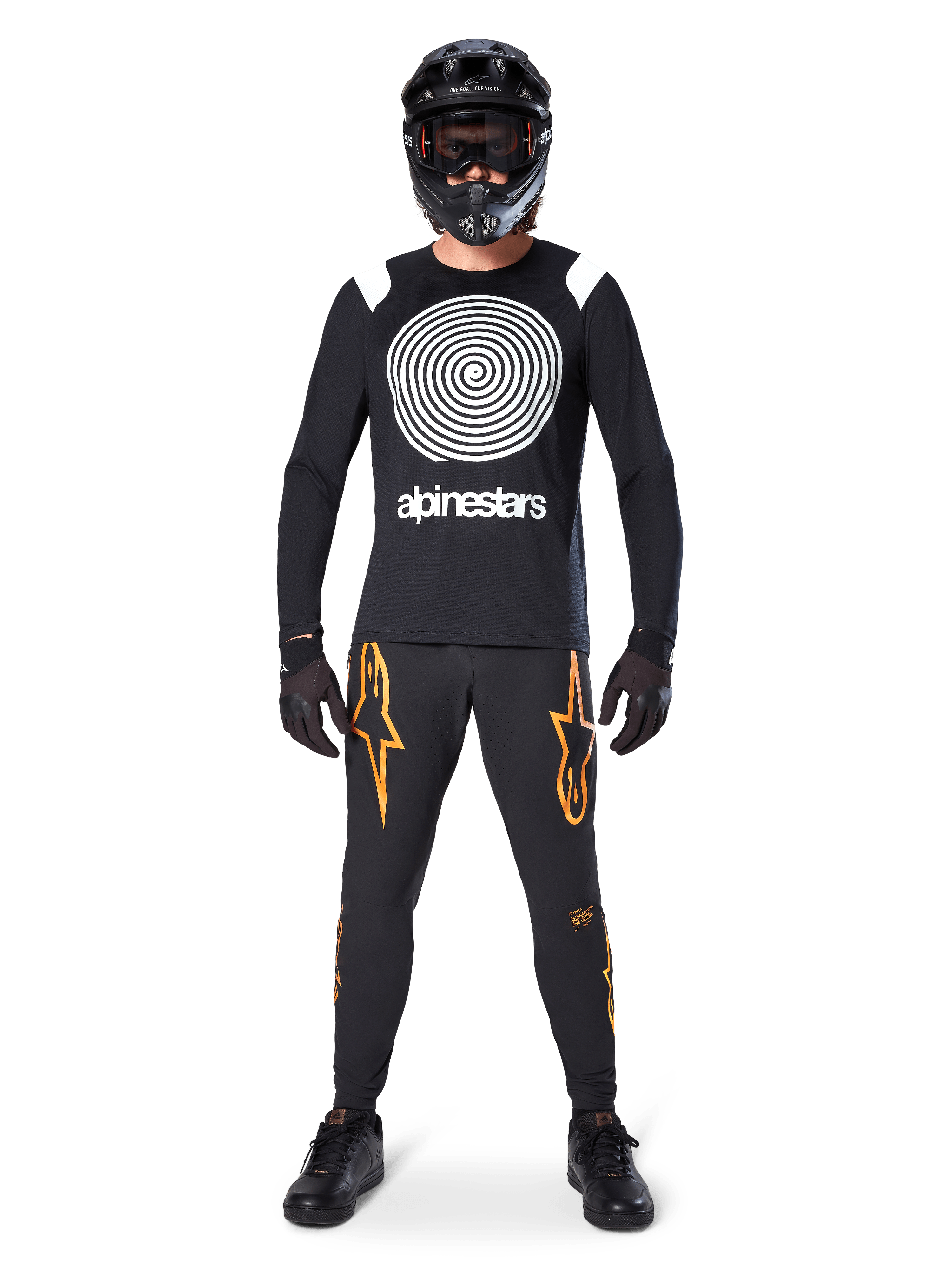 Mandlig model iført komplet mountainbike-udstyr, Alpinestars A-Supra Oscar langærmet trøje i sort med hvid spiralgrafik på brystet, sorte bukser med orange stjernelogoer, sort full-face hjelm og goggles, sorte handsker og sko, koordineret outfit til MTB gravity-kørsel