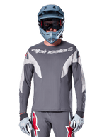 MTB-rytter iført Alpinestars A-Supra Race Jersey - Long Sleeve i Dark Gray, langærmet cykeltrøje med hvidt Alpinestars-logo, kombineret med blågrå full-face hjelm, goggles og grå racinghandsker, komplet mountainbike-racerudstyr til off-road performance