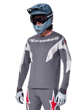 Alpinestars A-Supra Race Jersey - langærmet, mørkegrå med hvide og røde accenter, grafik i racing-stil, båret med matchende grå bukser, blå full-face hjelm og hvide handsker, professionelt MTB- og downhill-racerudstyr