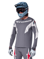 Alpinestars A-Supra Race Jersey - langærmet, mørkegrå med hvide og røde accenter, grafik i racing-stil, båret med matchende grå bukser, blå full-face hjelm og hvide handsker, professionelt MTB- og downhill-racerudstyr