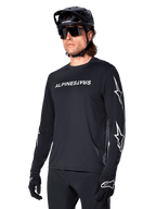 Male cyclist wearing Alpinestars A-Dura Switch Long Sleeve Jersey, Black with white wordmark and star logos on sleeves, styled with black MTB helmet and sunglasses, high-performance cycling gear for off-road riding