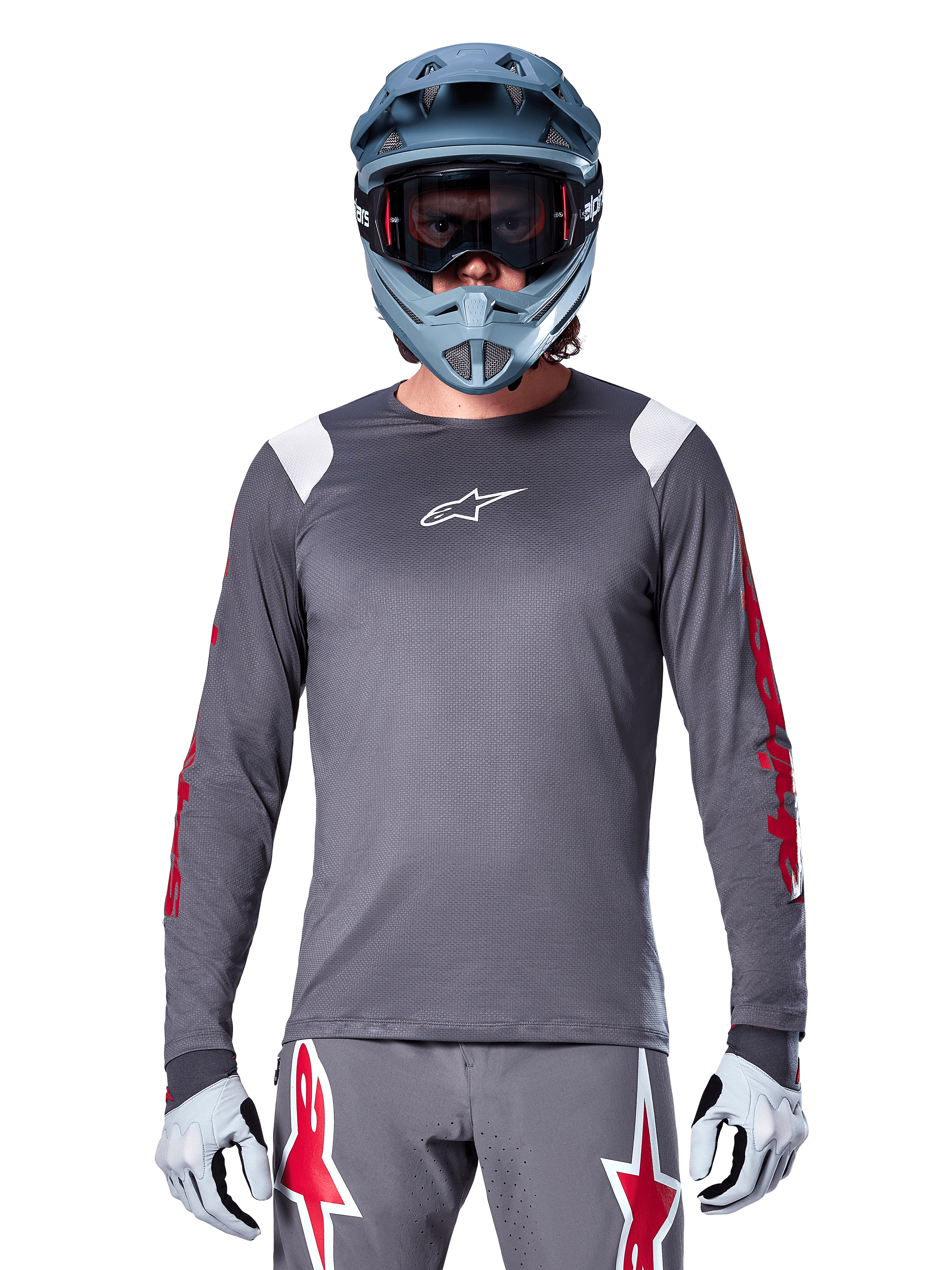 MTB rider wearing Alpinestars A-Supra Astar Long Sleeve Jersey, Dark Gray with red graphics on sleeves and white shoulder accents, matching gray pants with red star logos, blue full-face helmet and goggles, coordinated off-road cycling gear