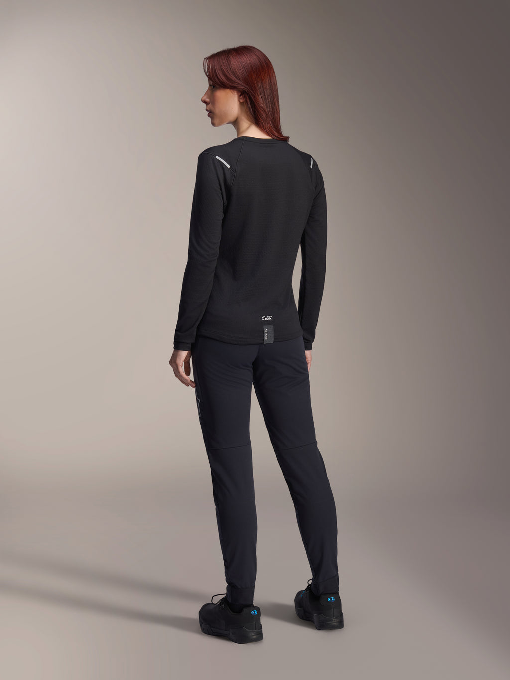 Alpinestars Stella A-Dura Thermal trøje til kvinder, langærmet MTB-trøje i sort, set bagfra med reflekterende skulderdetaljer og forlænget drop tail, kombineret med sorte bukser og sko, performance-udstyr til mountainbike.
