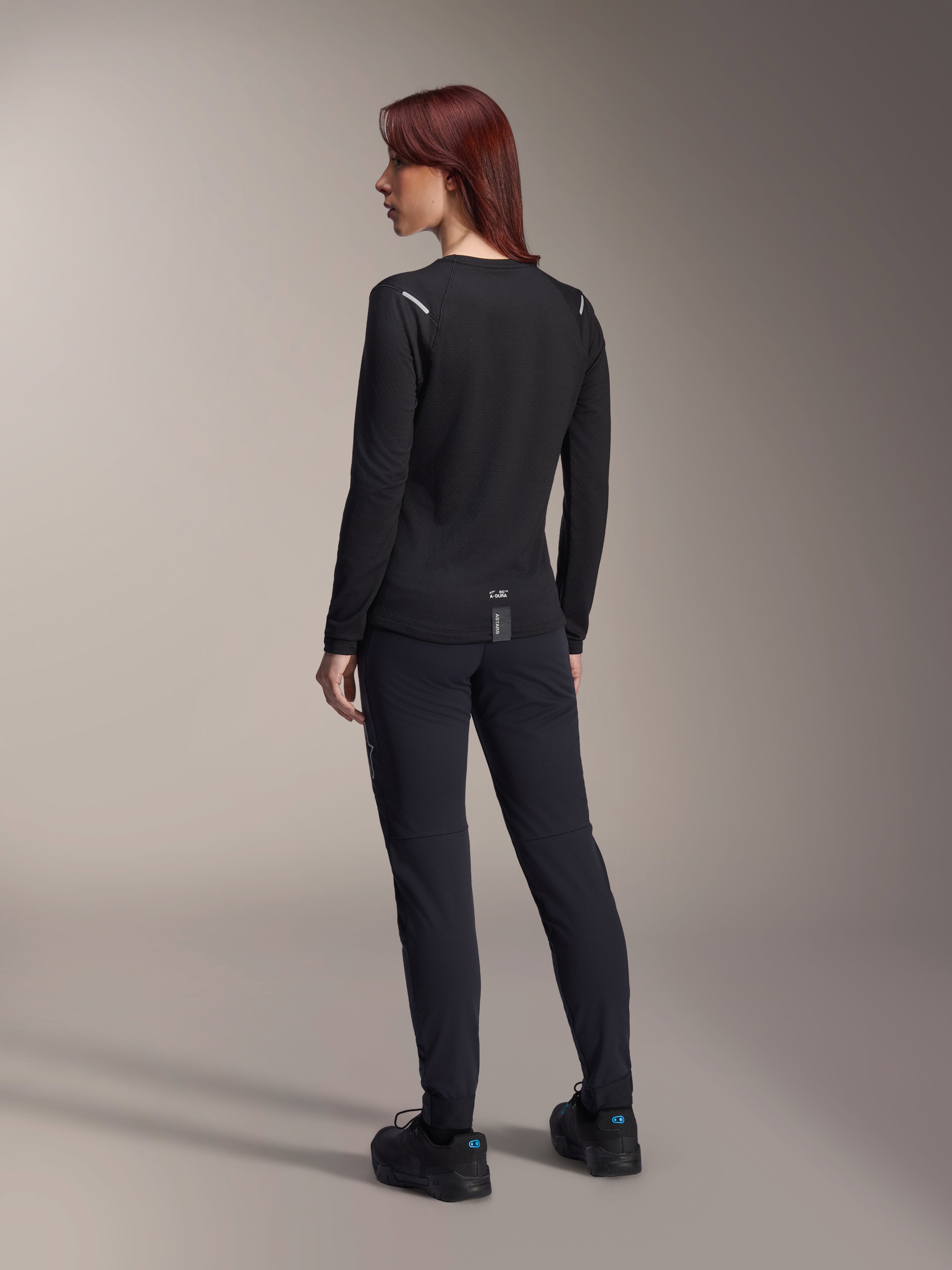 Alpinestars Stella A-Dura Thermal trøje til kvinder, langærmet MTB-trøje i sort, set bagfra med reflekterende skulderdetaljer og forlænget drop tail, kombineret med sorte bukser og sko, performance-udstyr til mountainbike.