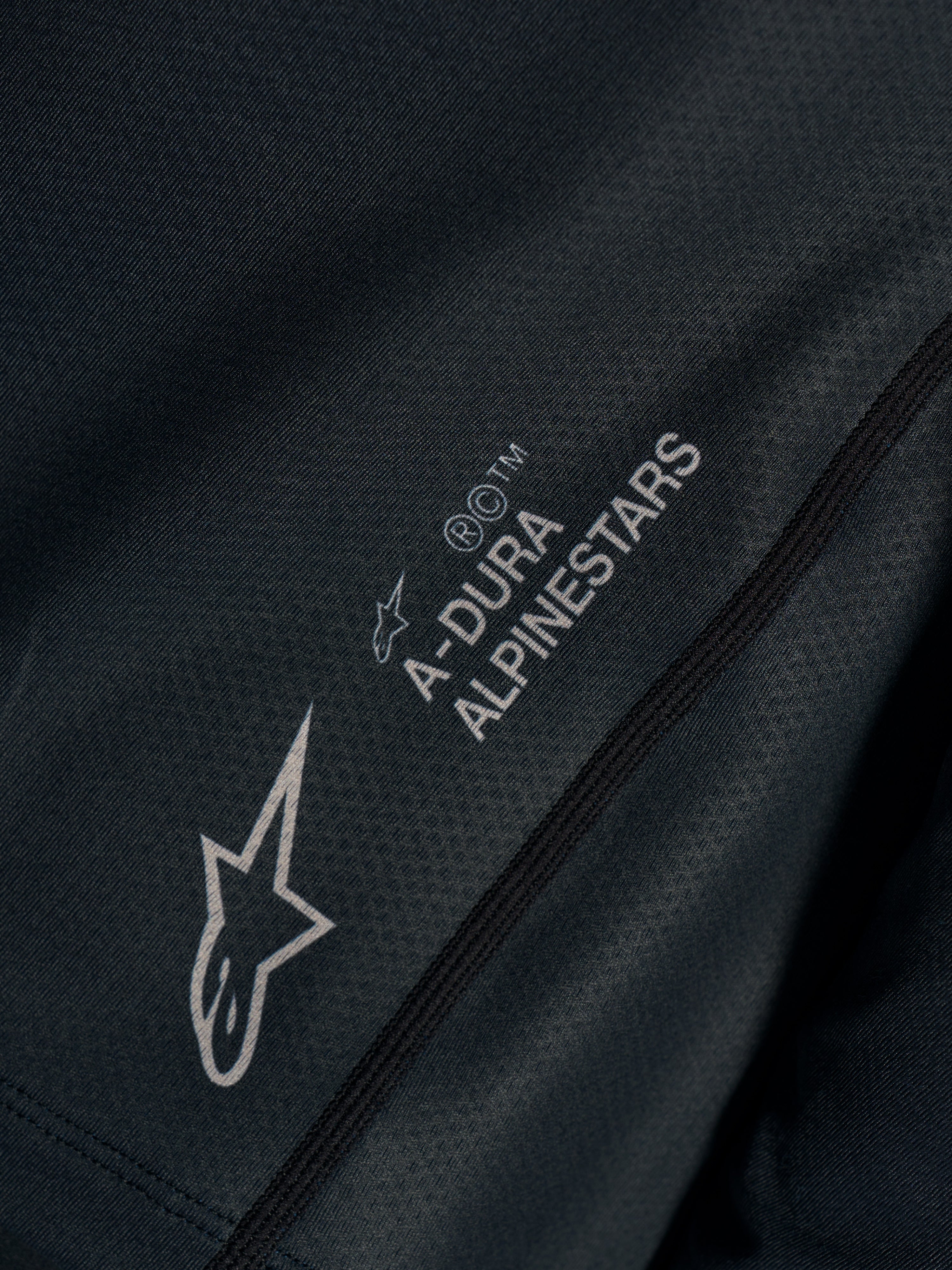Sort Stella A-Dura Ride trøje, nærbillede af teknisk jacquard-stof med A-DURA ALPINESTARS og Astars-logoer.