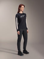Kvinde iført Alpinestars MTB-udstyr, sort Stella A-Dura Elite Jersey med hvidt brystlogo og hvide ærmepaneler, sorte Stella A-Dura bukser, sorte cykelsko med blå detaljer, komplet performance-outfit til mountainbiking