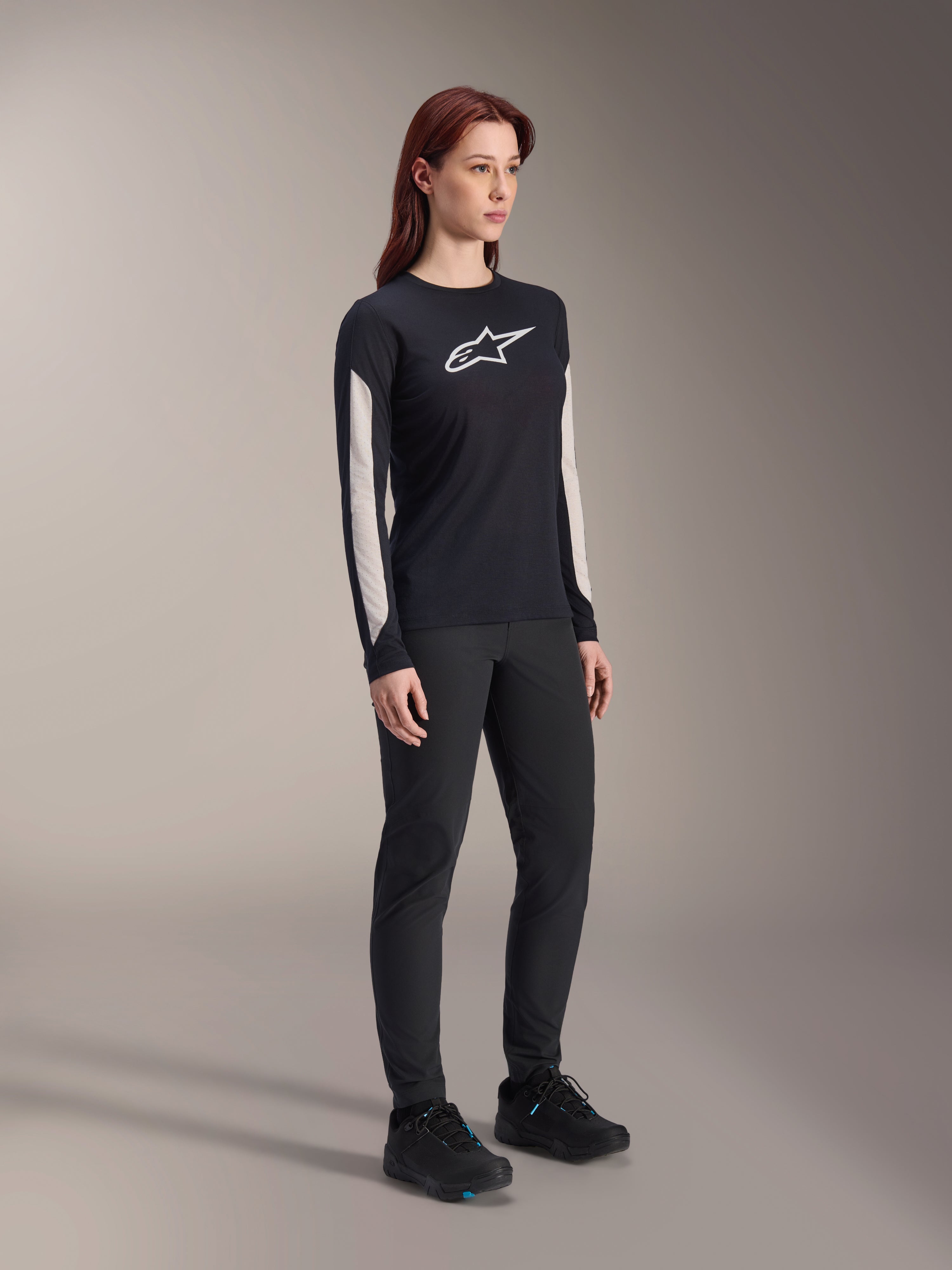 Kvinde iført Alpinestars MTB-udstyr, sort Stella A-Dura Elite Jersey med hvidt brystlogo og hvide ærmepaneler, sorte Stella A-Dura bukser, sorte cykelsko med blå detaljer, komplet performance-outfit til mountainbiking