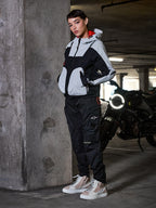 Alpinestars Actea damebukser, sorte, kvindelig motorcyklist iført motorcykelbukser i cargo-stil med en grå og sort teknisk jakke og beige sneakers, urbane omgivelser i en parkeringskælder med motorcykel.