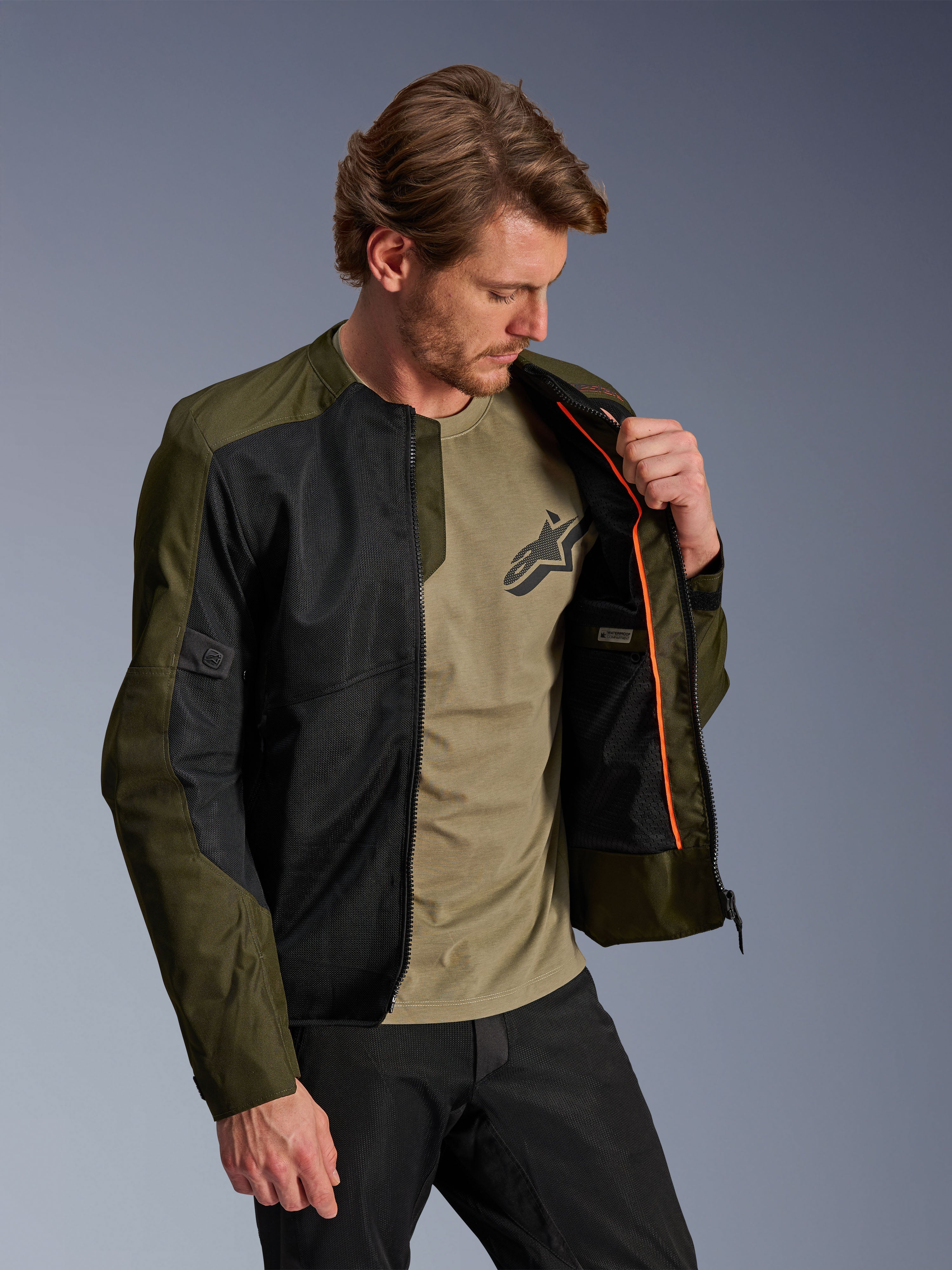 Alpinestars C-1 Air-jakke i khaki og sort, båret af en mandlig model for at vise det orange inderfor og det vandtætte rum, store tech mesh-paneler til luftgennemstrømning, urban moderne motorcykelstil kombineret med en matchende oliven t-shirt og sorte bukser