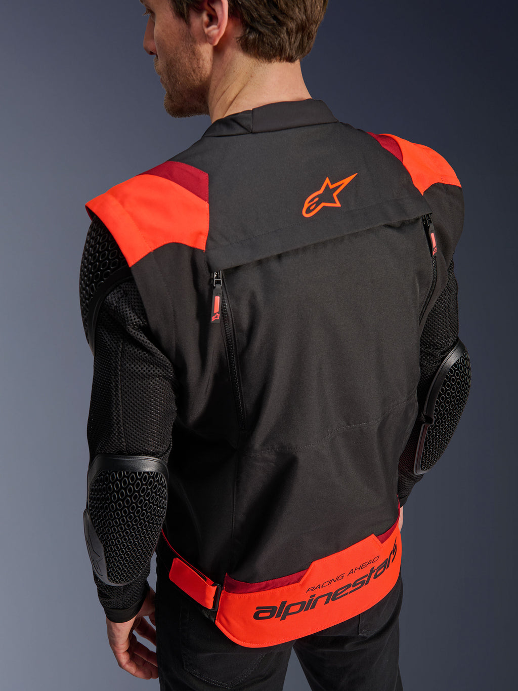 Alpinestars T-Stunt Air-jakke, sort, rød fluo og mørkerød, set bagfra på en model, viser mesh-ærmer med bikubestrukturerede albuebeskyttere, Alpinestars-stjernelogo på den øvre ryg og markant branding på den nedre ryg med 'RACING AHEAD'-tekst, modulært sportssnit-design til motorcykelkørsel.