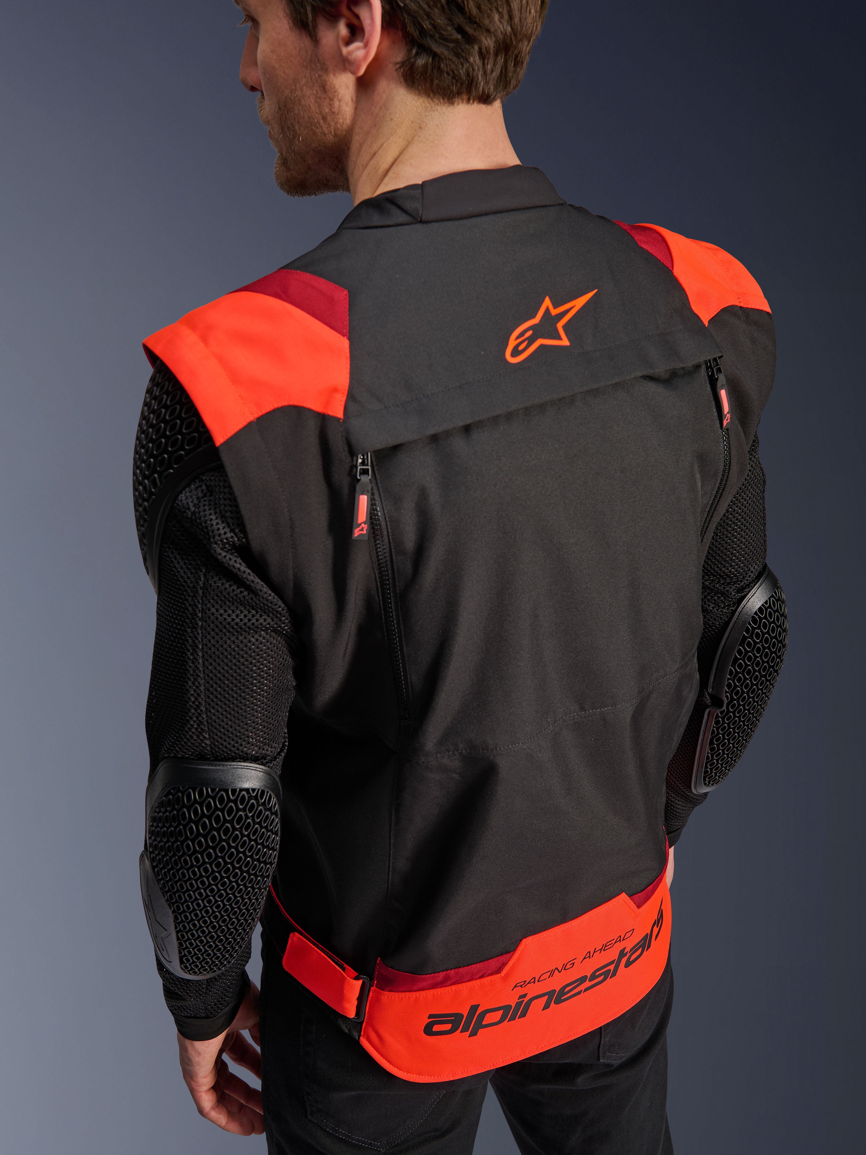 Alpinestars T-Stunt Air-jakke, sort, rød fluo og mørkerød, set bagfra på en model, viser mesh-ærmer med bikubestrukturerede albuebeskyttere, Alpinestars-stjernelogo på den øvre ryg og markant branding på den nedre ryg med 'RACING AHEAD'-tekst, modulært sportssnit-design til motorcykelkørsel.
