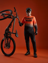 A-Dura ASTARS Jersey 2.0 langærmet trøje i orange-sort gradient med brystlogo, vist med komplet mountainbike-udstyr.