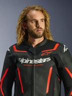 Alpinestars GP Force V2 læderjakke i sort og rød fluo, båret af en mandlig model med langt krøllet blond hår, med markant hvid Alpinestars-branding og 'RACING AHEAD'-tekst på brystet, premium motorcykeljakke i læder designet til sportskørsel.