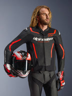 Alpinestars GP Force V2 læderjakke, sort og rød fluo, båret af en mandlig model, der holder en matchende hjelm, med hvide Alpinestars-logoer og røde detaljer, kombineret med læderbukser og racerhandsker, sportsmotorcykeludstyr til vej og bane.