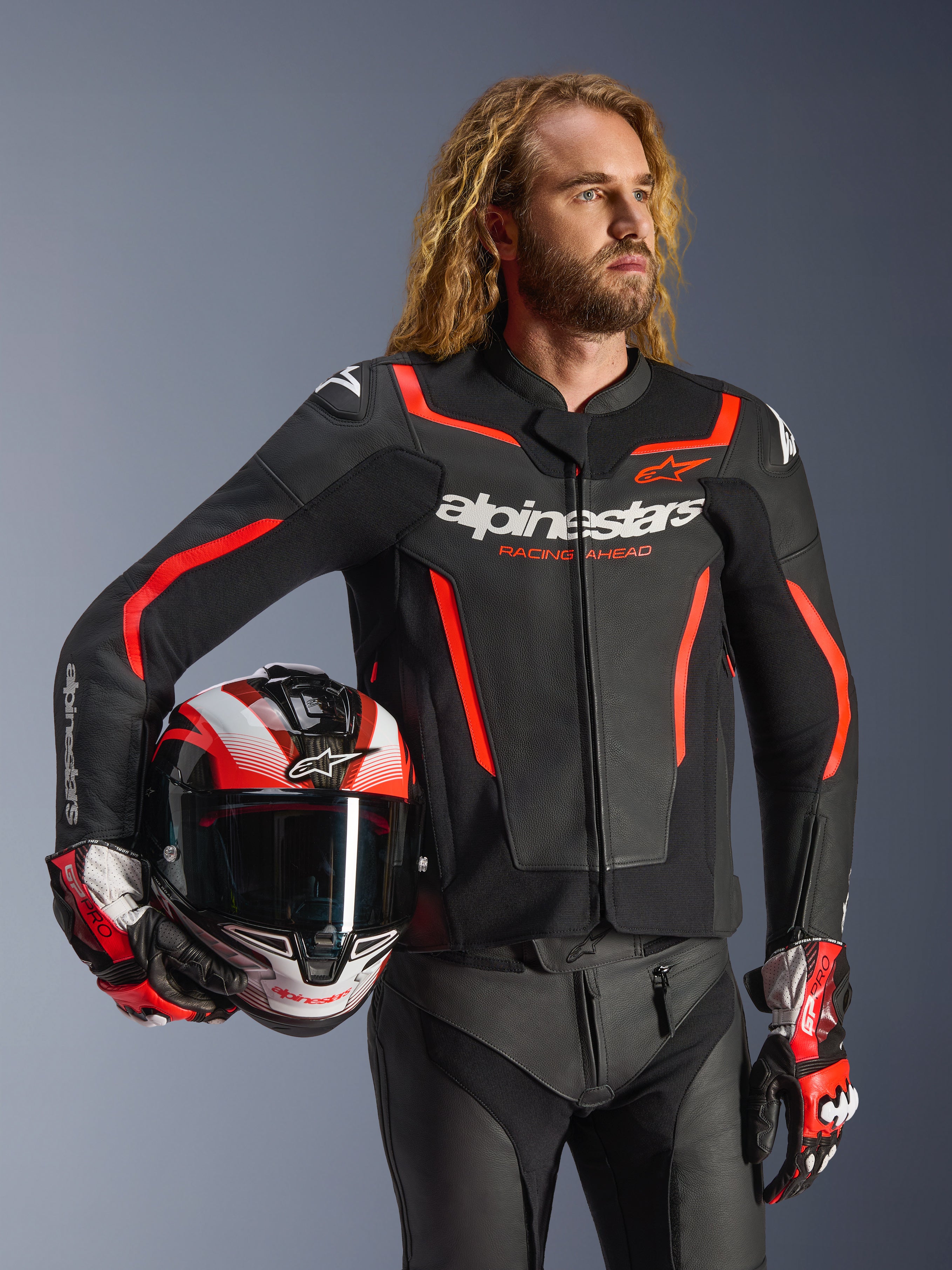 Alpinestars GP Force V2 læderjakke, sort og rød fluo, båret af en mandlig model, der holder en matchende hjelm, med hvide Alpinestars-logoer og røde detaljer, kombineret med læderbukser og racerhandsker, sportsmotorcykeludstyr til vej og bane.