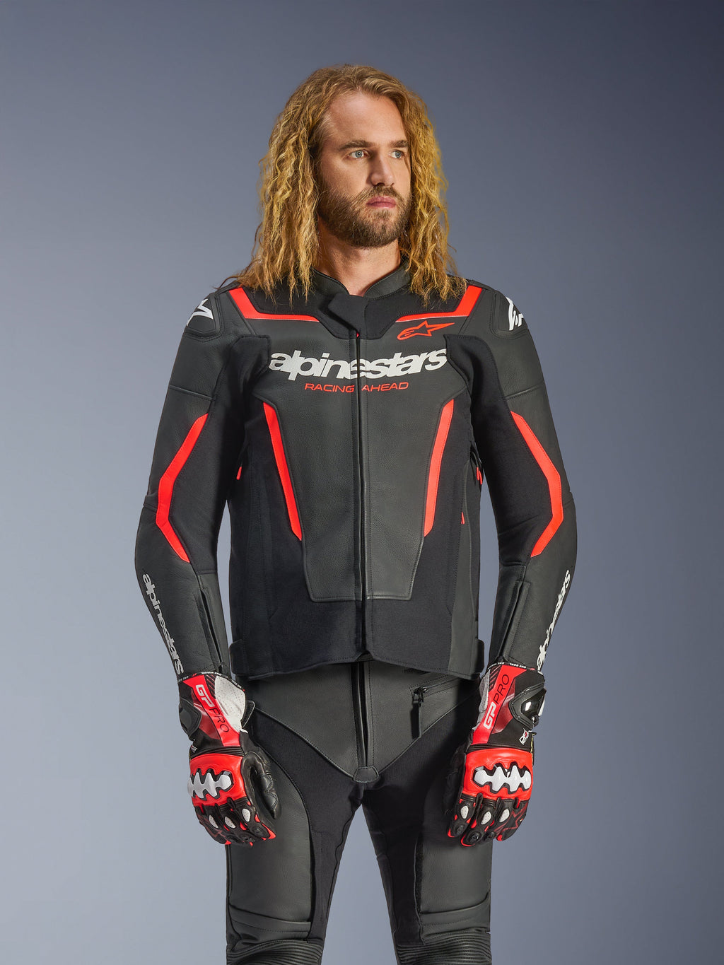 Alpinestars GP Force V2 læderjakke, sportsmotorcykeljakke, sort og rød fluo, båret af en mandlig model med langt bølget hår, med hvidt Alpinestars-logo og "RACING AHEAD"-tekst, kombineret med matchende læderbukser og handsker, professionelt køreudstyr