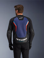 Set bagfra af Alpinestars GP Force V2 læderjakken i Black, Blue og Red Fluo, med et stort blåt rygpanel med fluorescerende røde og lyseblå detaljer, hvidt Alpinestars-logo og 'Racing Ahead'-branding på den nederste del af ryggen, båret af en kører med sorte handsker og bukser.
