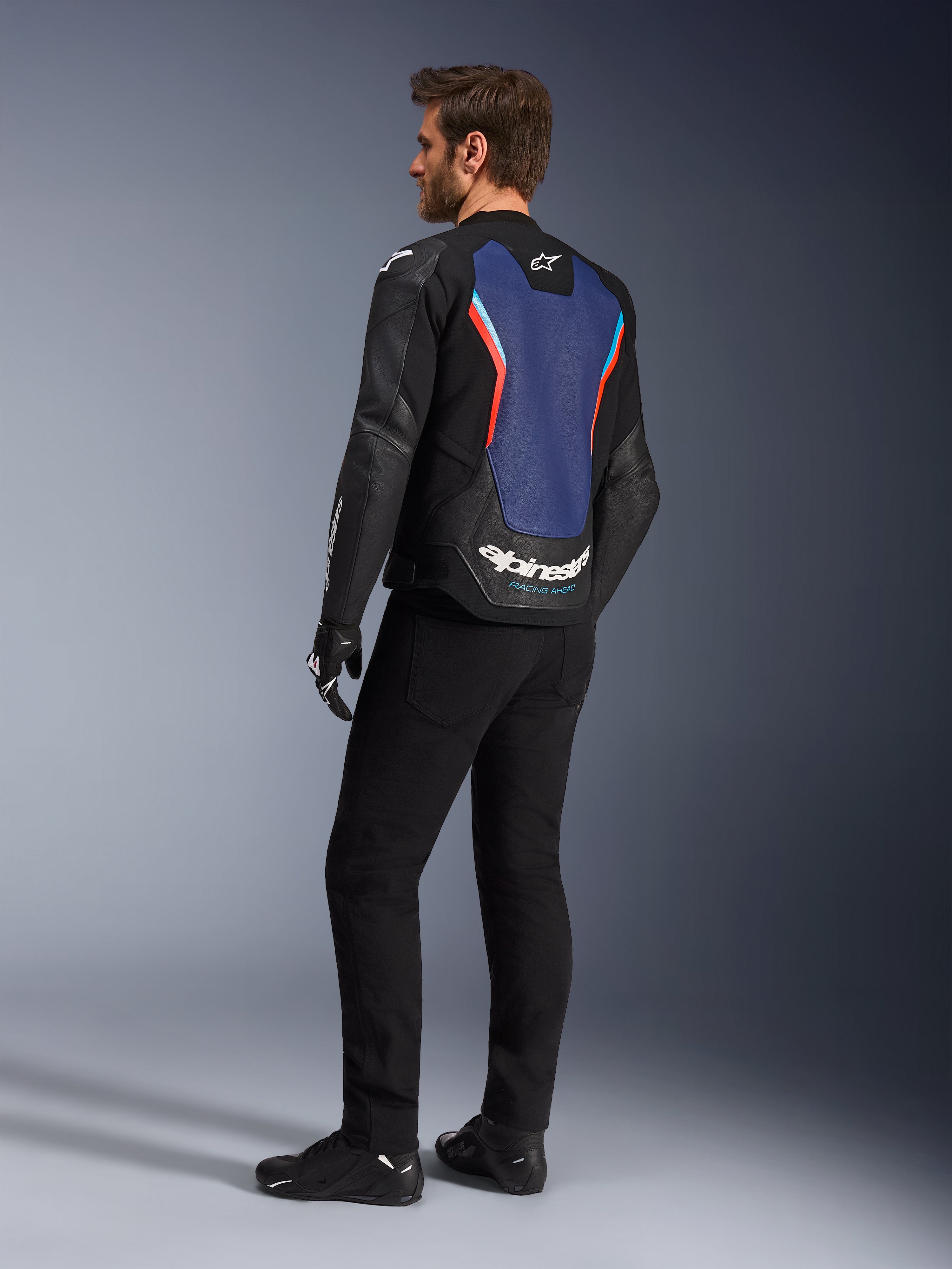 Trekvart profil bagfra af en mandlig model iført Alpinestars GP Force V2 læderjakke i Black Blue Red Fluo, der fremhæver det blå rygpanel med fluorescerende røde detaljer og hvide logoer, stylet med sorte bukser og motorcykelhandsker.