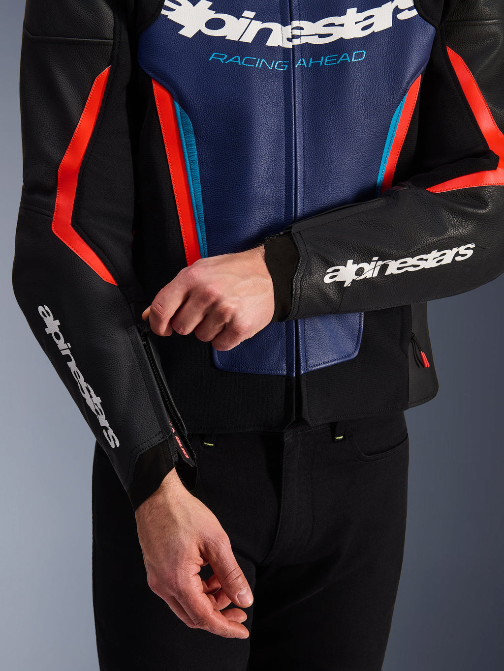 Alpinestars GP Force V2 læderjakke i sort, blå og rød fluo, nærbillede af en person, der justerer lynlåsen ved ærmemanchetten, med hvide Alpinestars-logoer på underarmen og brystet, tekstureret okselæderkonstruktion til sportslig motorcykelkørsel