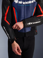 Alpinestars GP Force V2 læderjakke i sort, blå og rød fluo, nærbillede af en person, der justerer lynlåsen ved ærmemanchetten, med hvide Alpinestars-logoer på underarmen og brystet, tekstureret okselæderkonstruktion til sportslig motorcykelkørsel