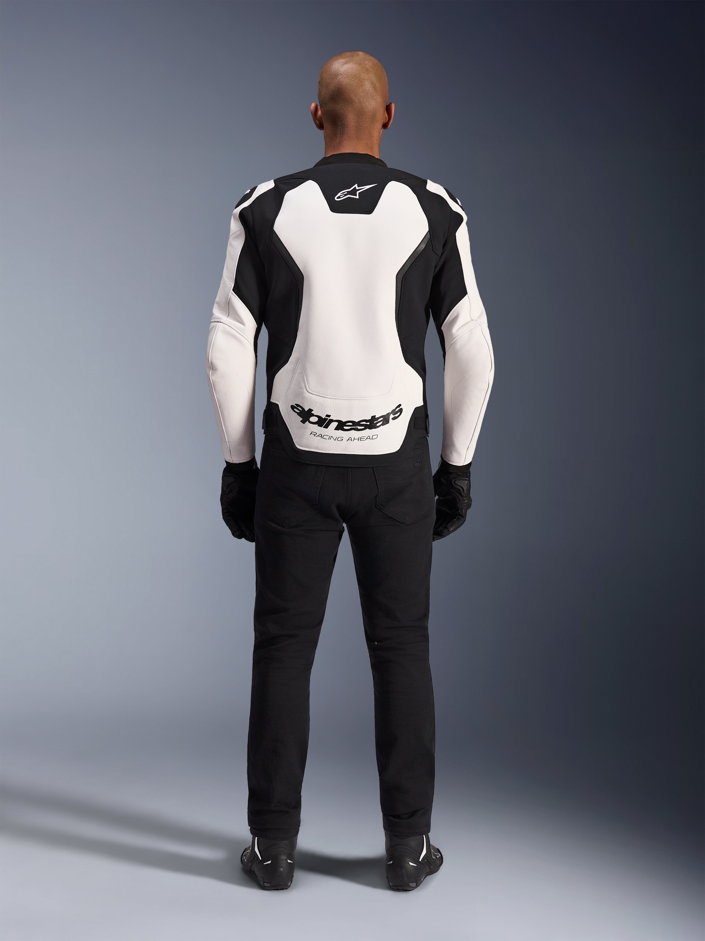 Alpinestars GP Force V2 Leather Jacket, White and Black, rear view showing large white back panel with Alpinestars logo and 'RACING AHEAD' text, worn with black riding pants and gloves, sport motorcycle gear design