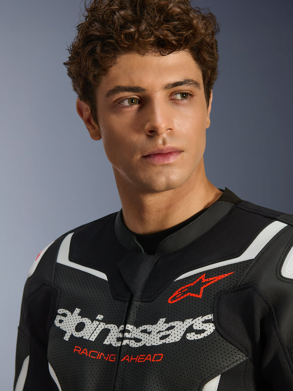 Portræt af en mandlig model iført Alpinestars GP Force V2 Air læderjakke i sort og hvid, med perforeret læderkonstruktion, fremtrædende hvidt logo og rød 'RACING AHEAD'-tekst på brystet, sportsmotorcykeludstyr