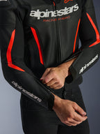Alpinestars GP Force V2 Air læderjakke i sort og rød fluo, nærbillede der viser den perforerede læderkonstruktion, hvidt branding på bryst og ærmer, og en kører, der justerer lynlåsen på underarmen for en skræddersyet pasform.