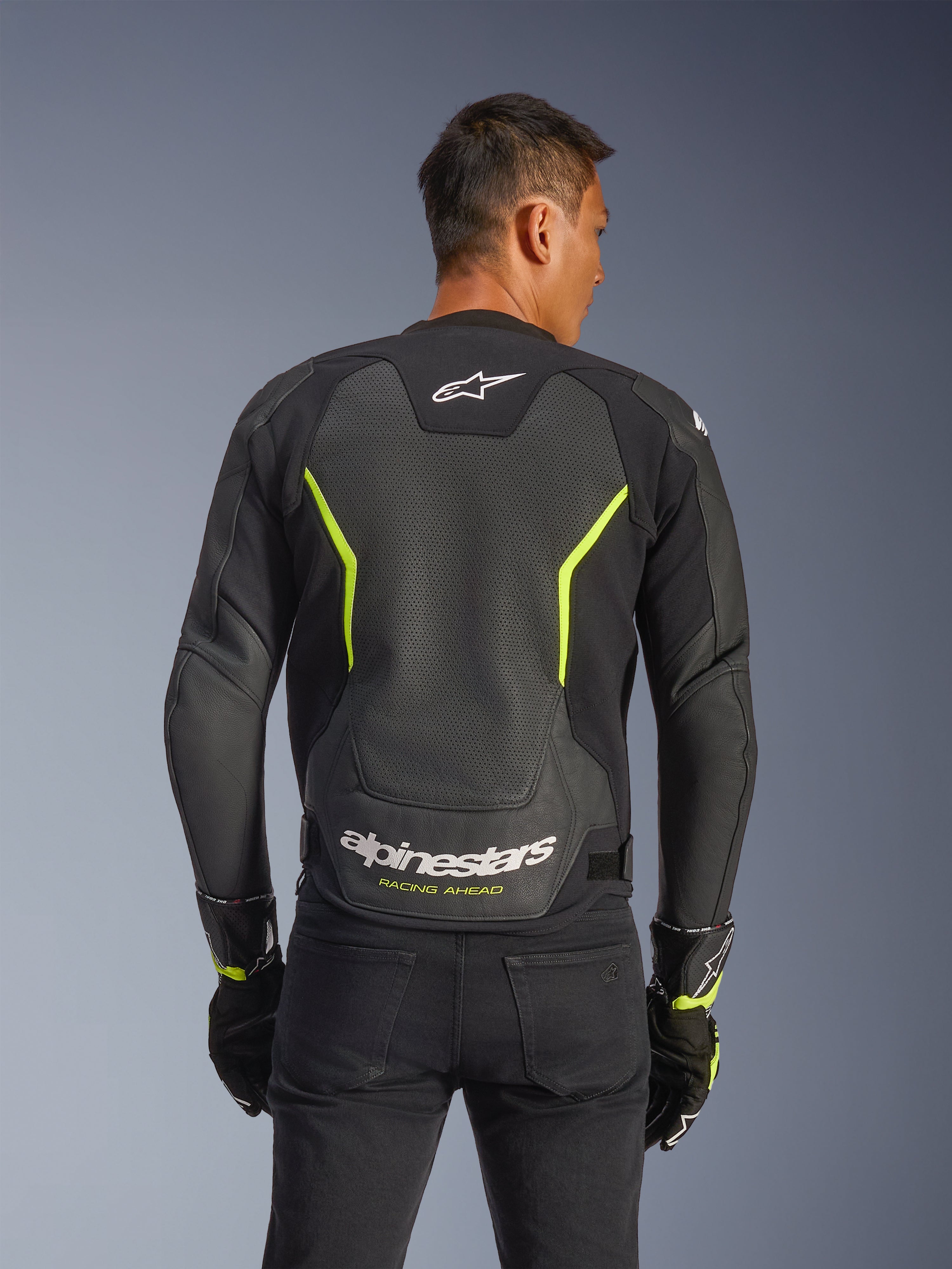 Alpinestars GP Force V2 Air læderjakke, sort og gul fluo, rygvisning af en mandlig kører med omfattende perforerede læderpaneler for luftstrøm, hvid Alpinestars-branding og gul "Racing Ahead"-tekst på den nederste del af ryggen, sportsmotorcykeludstyr til vejbrug.
