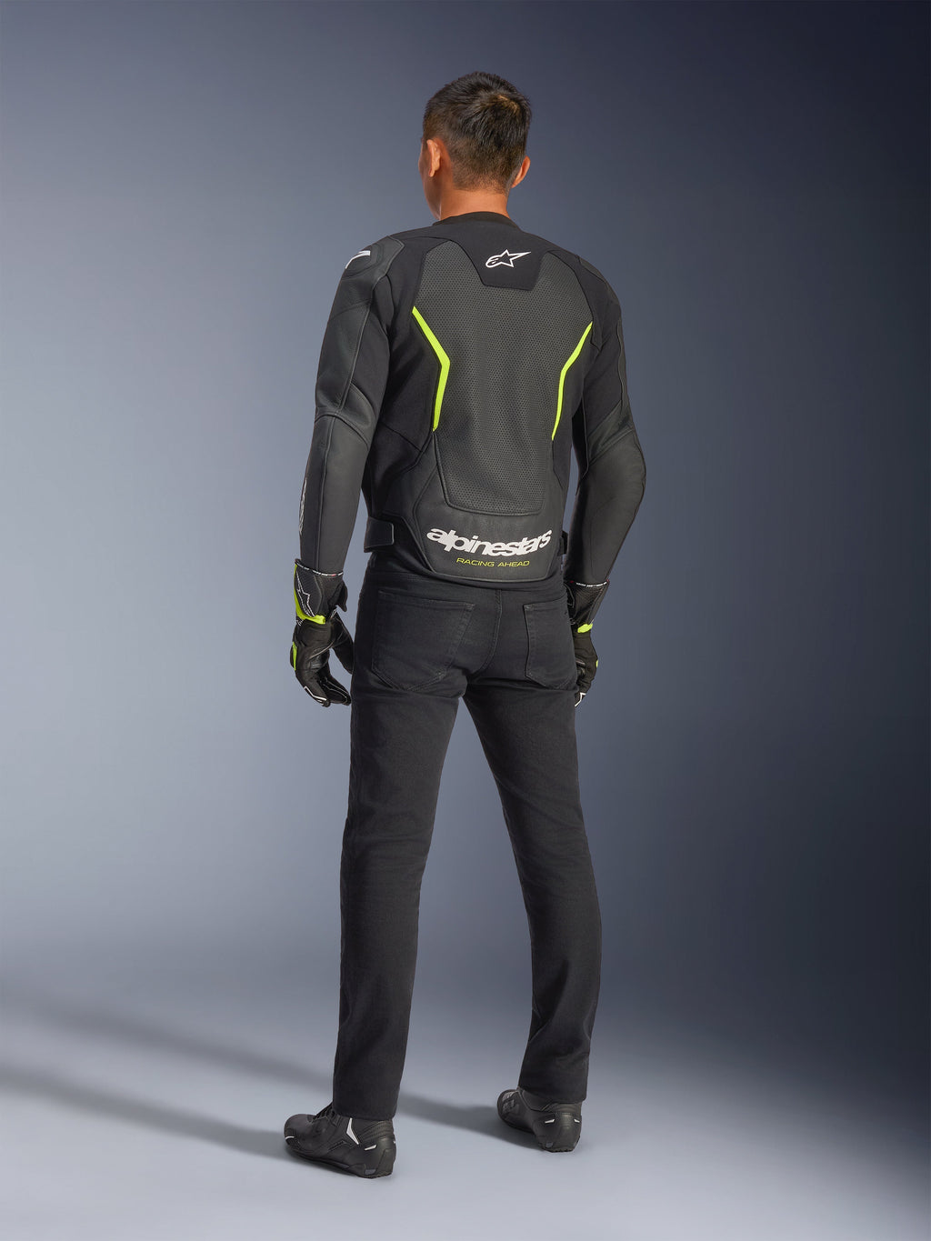 Alpinestars GP Force V2 Air læderjakke, sort og gul fluo, rygvisning af en kører iført den perforerede læderjakke, med vinklede gule detaljer og hvidt Alpinestars-logo med 'RACING AHEAD' på den nederste del af ryggen, kombineret med sorte kørebukser og støvler.