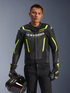 Mandlig motorcyklist iført Alpinestars GP Force V2 Air læderjakke i sort og gul fluo, holder en sort motorcykelhjelm, perforeret læder med hvid Alpinestars-branding og neon gule detaljer, matchende racerhandsker og sorte bukser