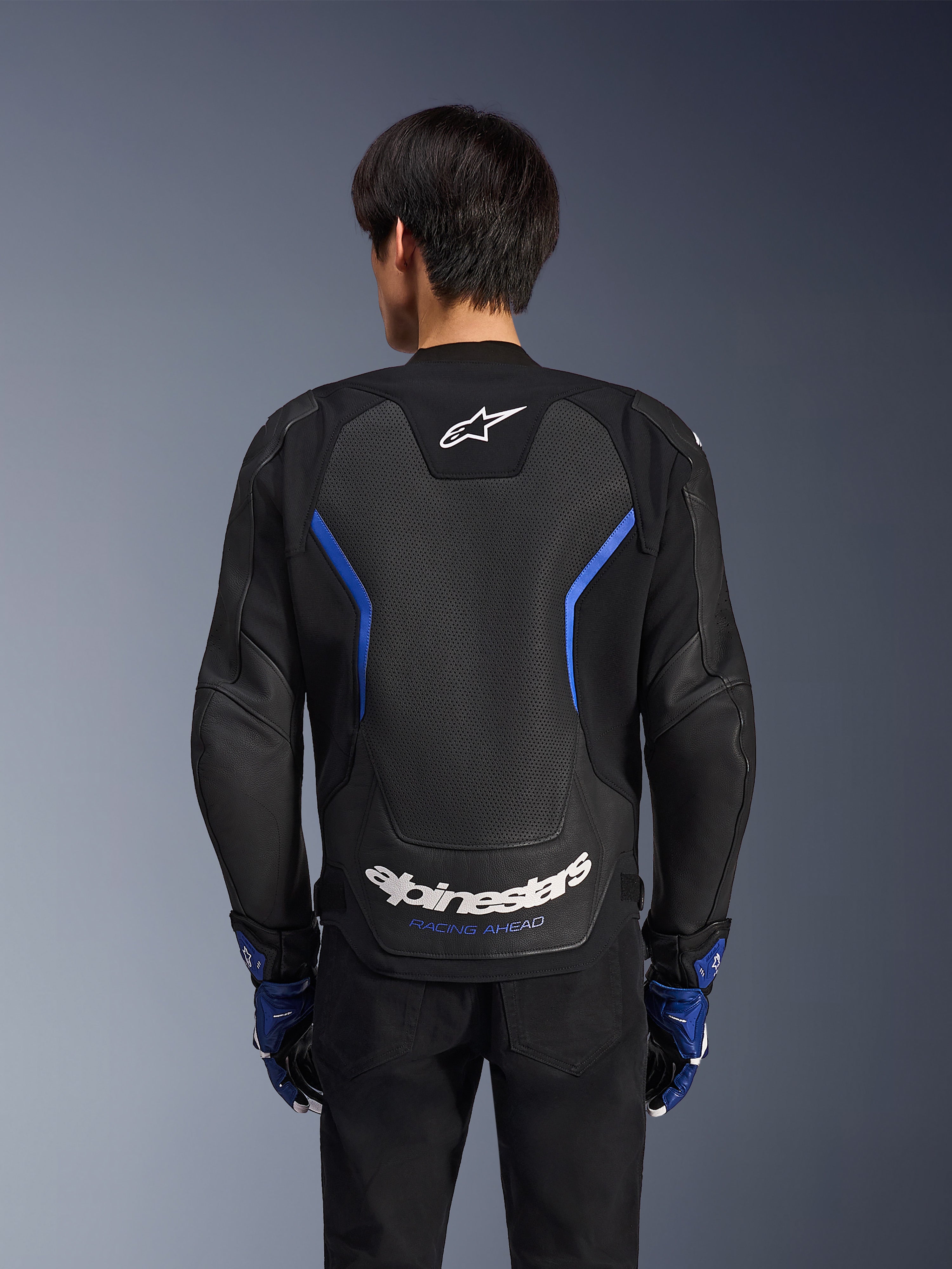 Alpinestars GP Force V2 Airflow læderjakke, Sort Metallic Blå, set bagfra, med et stort perforeret rygpanel til ventilation, blå detaljer og hvid Alpinestars-branding, båret med matchende blå handsker og sorte bukser til sportskørsel.