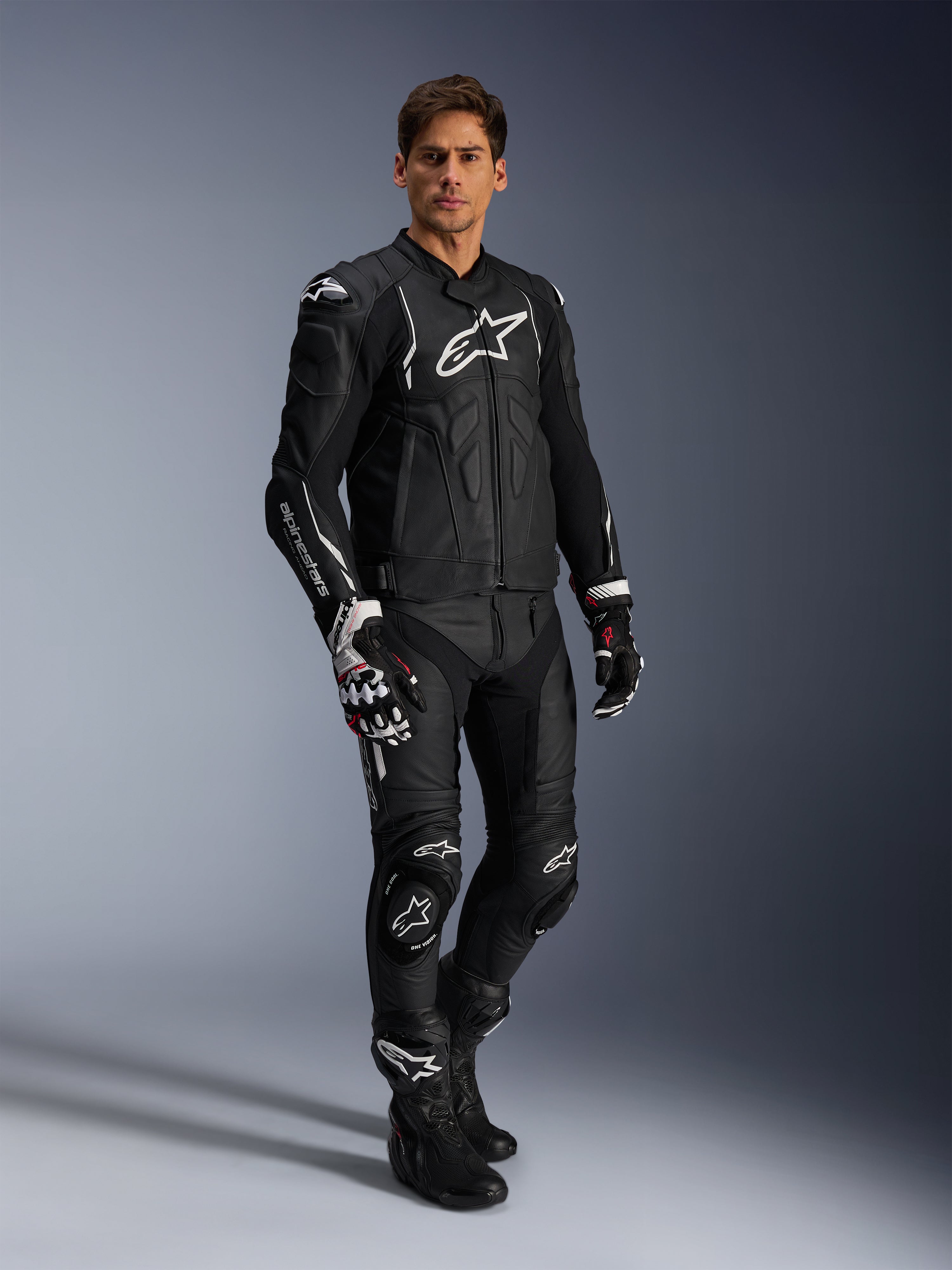 Mandlig motorcyklist iført Alpinestars Dusk læderjakke i sort og hvid, premium læderjakke med stort hvidt logo på brystet, kombineret med matchende læderracerbukser, handsker og støvler, komplet performance sports-køreudstyr.