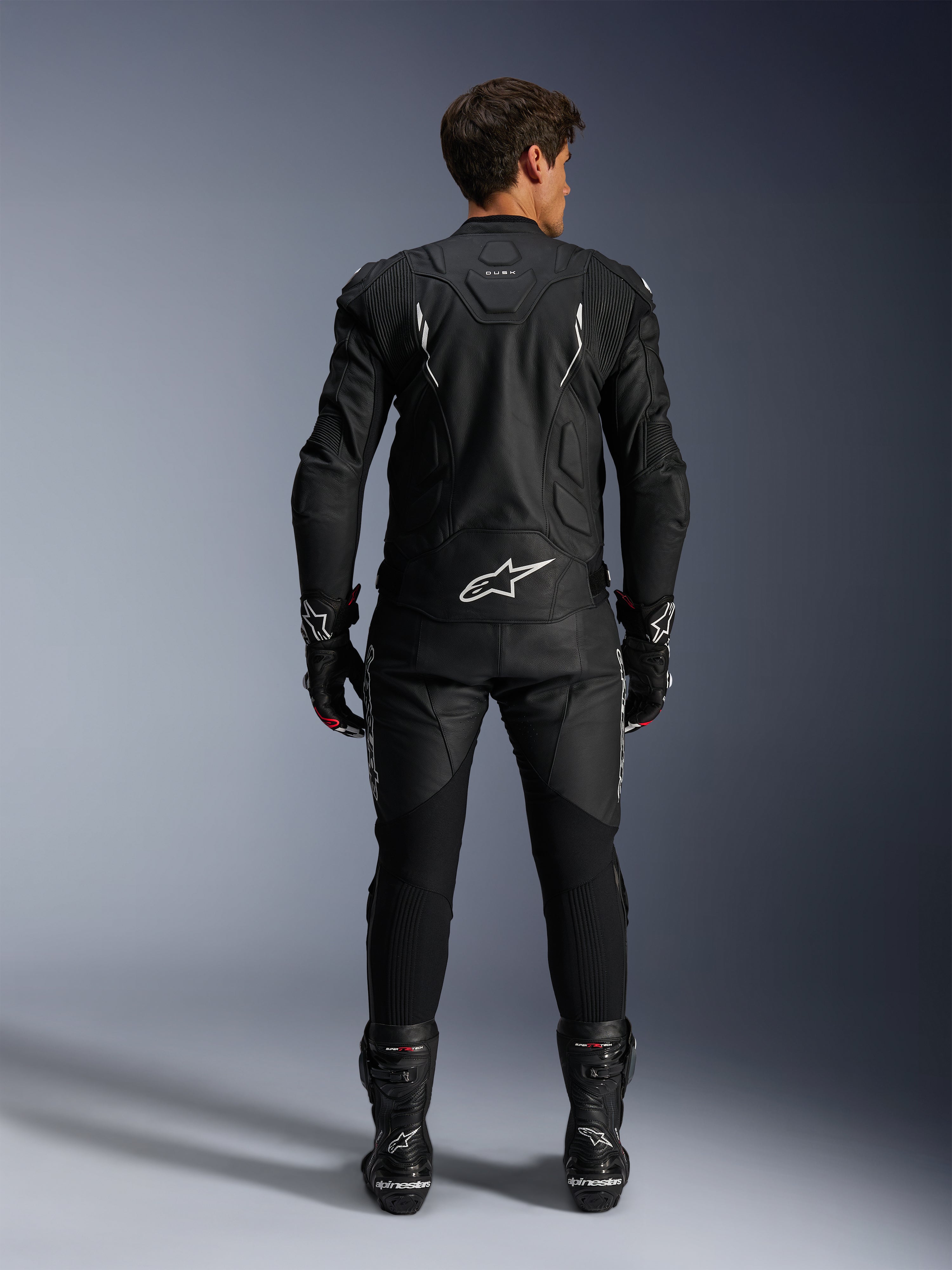 Motorcykelrytter set bagfra iført Alpinestars Dusk læderjakke og Missile V3 læderbukser, komplet sort læderdragt med hvide detaljer, jakken har 'Dusk'-branding og Alpinestars-logo på ryggen, kombineret med sorte racingstøvler og handsker, koordineret motorsport-outfit til vejkørsel.
