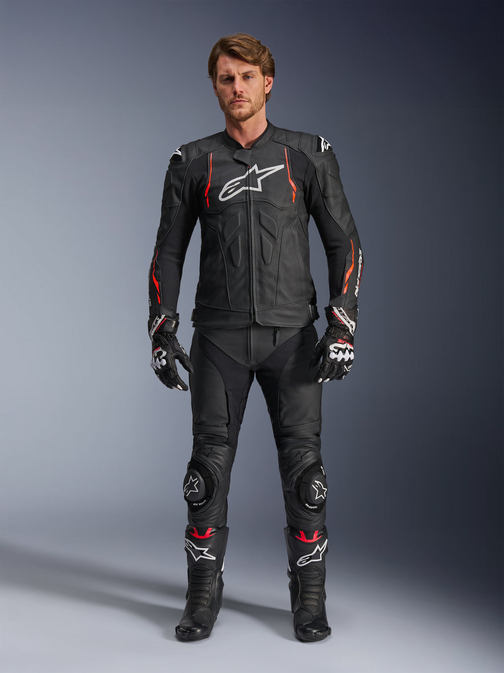 Mandlig motorcyklist iført Alpinestars Dusk Airflow læderjakke i Black Red Fluo, med perforerede læderpaneler og hvidt logo på brystet, kombineret med matchende sorte læder-kørebukser, knæslidere, handsker og racerstøvler, komplet motorcykeludstyr til vejbrug.