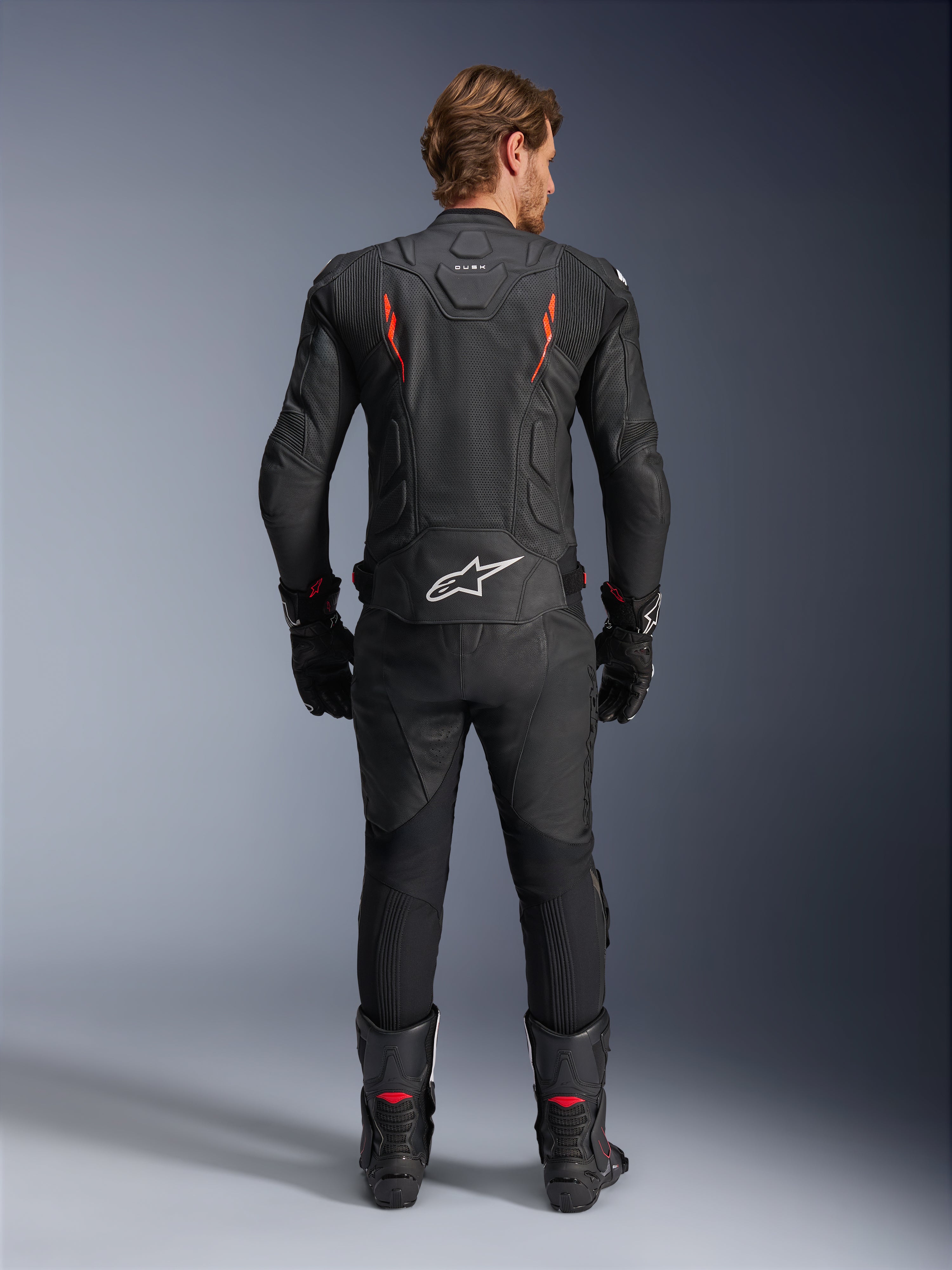 Alpinestars Dusk Airflow læderjakke, sort og rød fluo, bagsidevisning med omfattende perforeringer og stort hvidt logo på lænden, båret med matchende racerudstyr i læder inklusive bukser, støvler og handsker