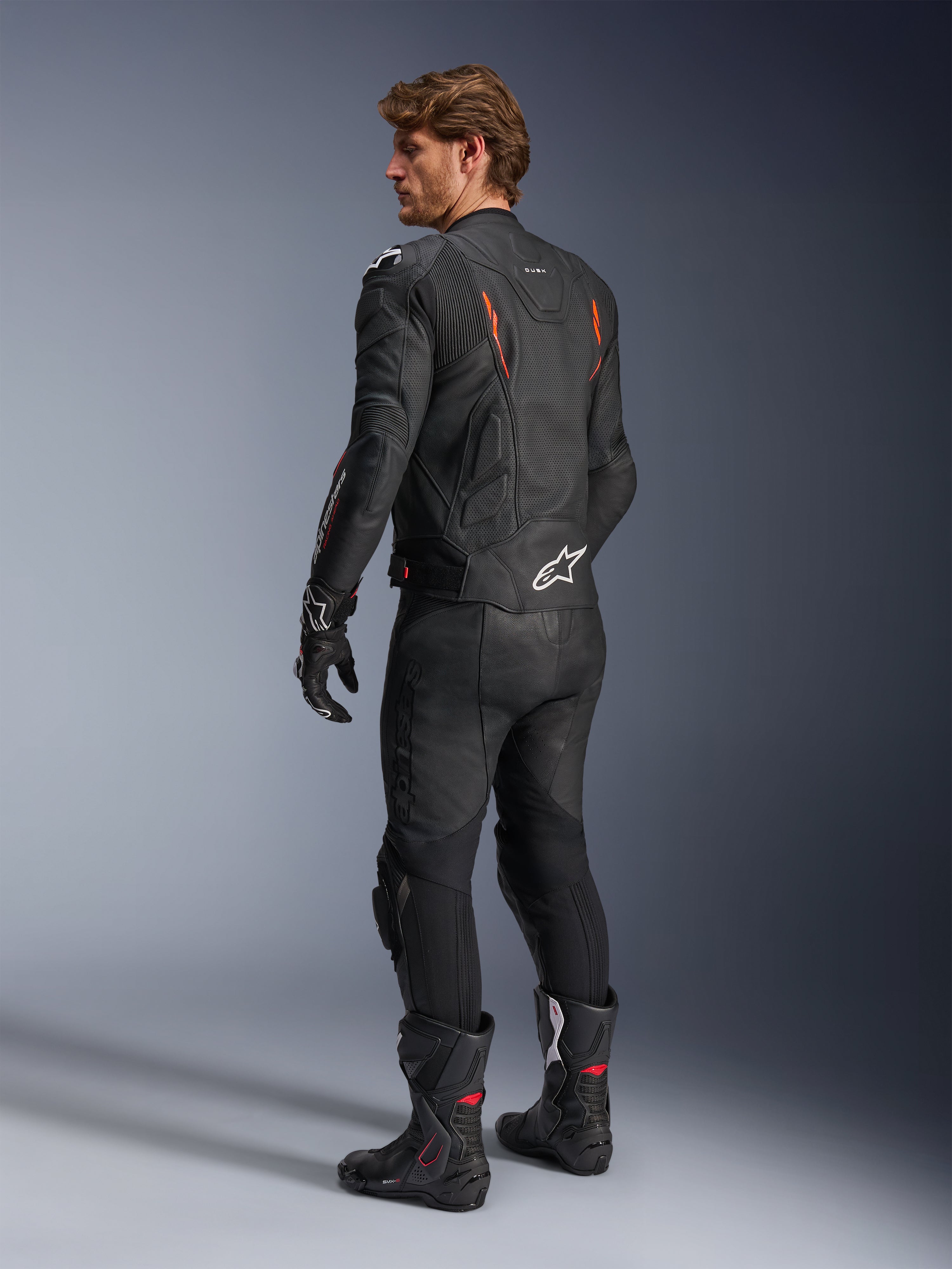 Alpinestars Dusk Airflow læderjakke, sort med Red Fluo-detaljer, set bagfra af en kører i fuldt lædermotorcykeludstyr inklusiv matchende sorte bukser, handsker og racerstøvler, med perforeret læder og hvide Alpinestars-logoer.