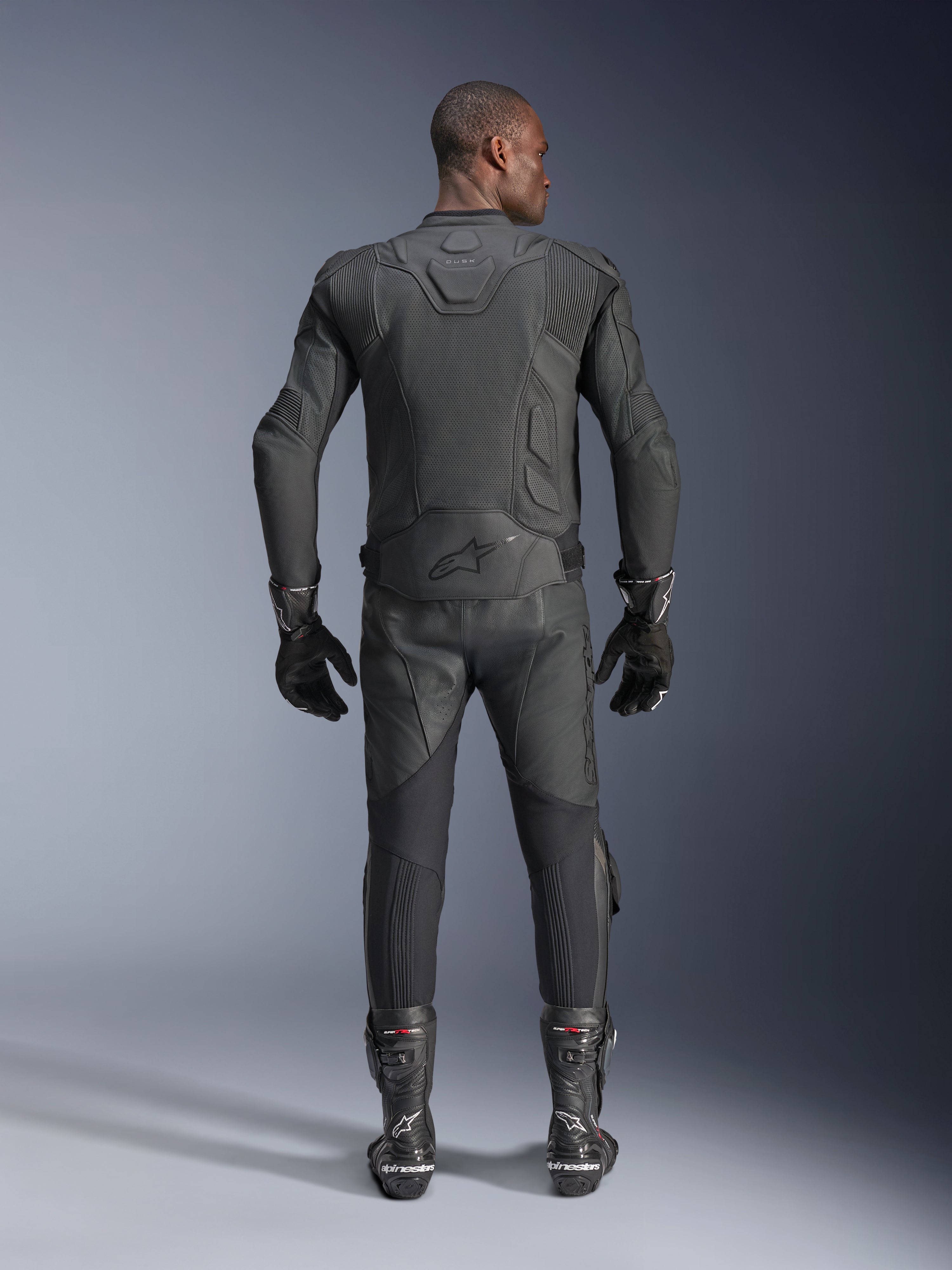 Mandlig model set bagfra iført Alpinestars Dusk Airflow læderjakke i sort, kombineret med matchende sorte læderracerbukser, handsker og støvler, med omfattende perforeringer for ventilation og Alpinestars-logo på lænden.
