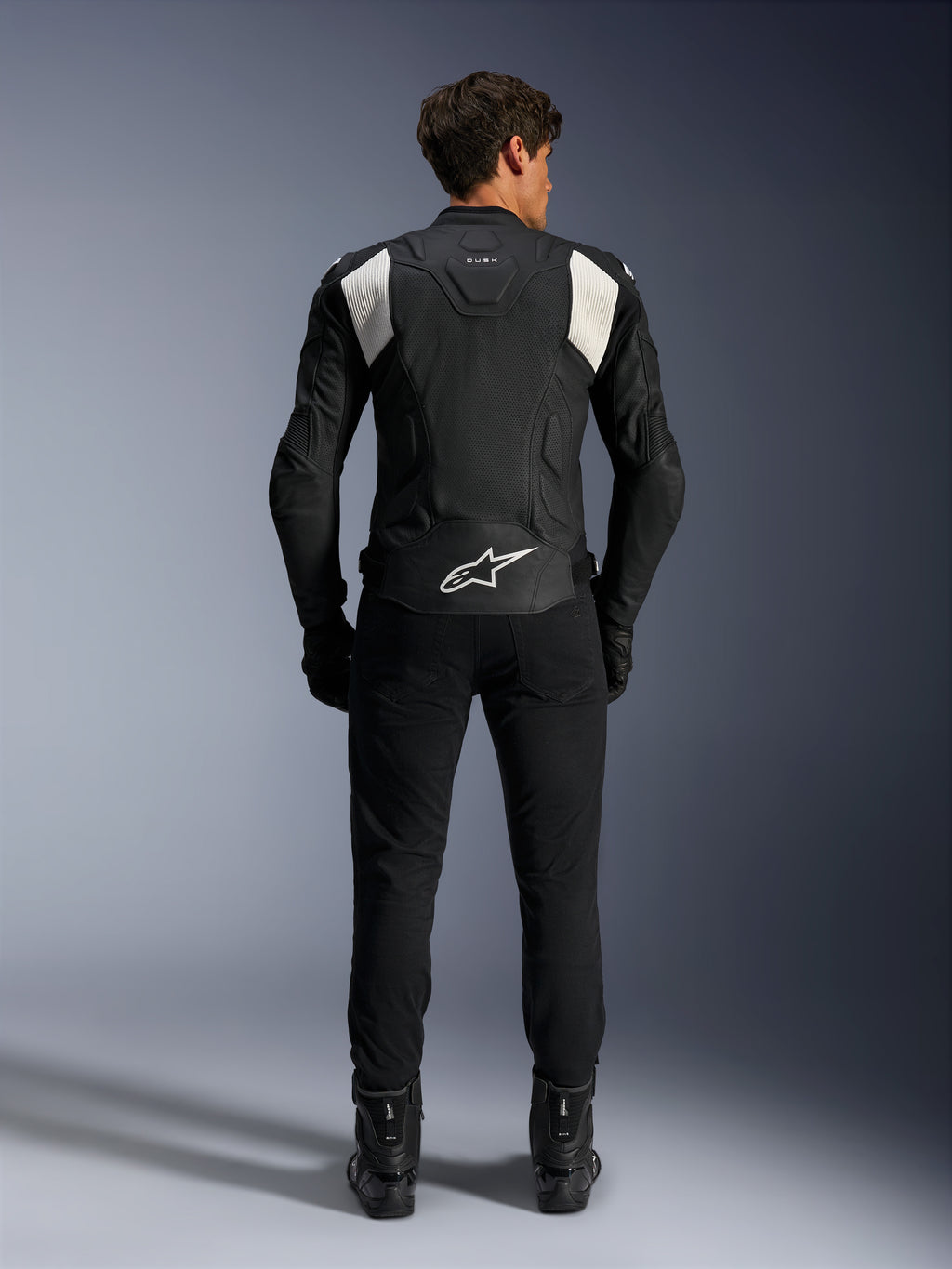 Alpinestars Dusk Airflow læderjakke, sort og hvid, set bagfra af en kører iført sorte bukser og støvler, med perforeret læder til ventilation, hvide accordion-strækpaneler på skuldrene og hvidt logo på den nederste del af ryggen