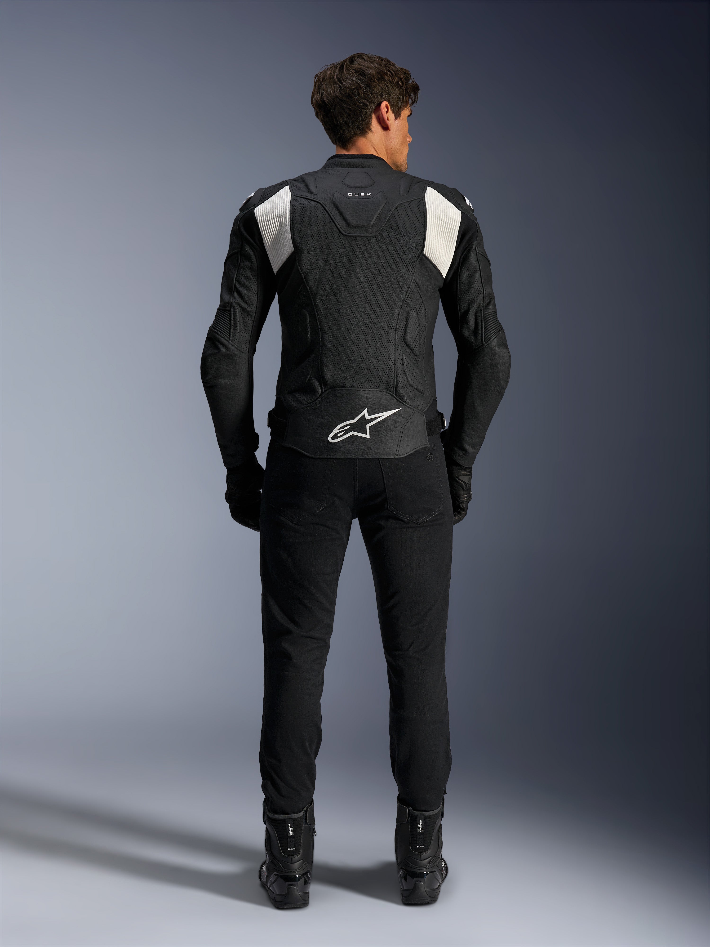 Alpinestars Dusk Airflow læderjakke, sort og hvid, set bagfra af en kører iført sorte bukser og støvler, med perforeret læder til ventilation, hvide accordion-strækpaneler på skuldrene og hvidt logo på den nederste del af ryggen